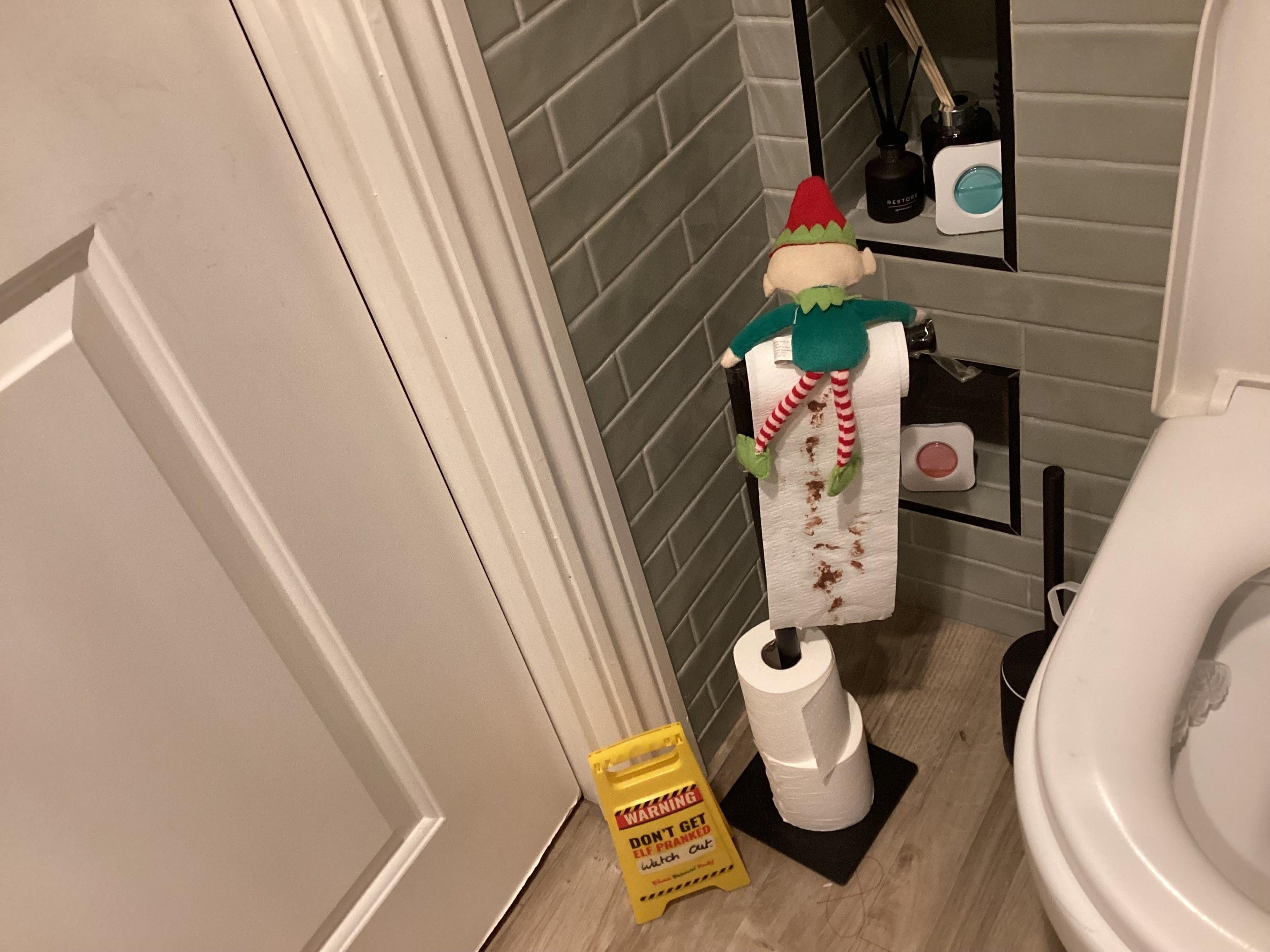 An elf is holding onto a toilet roll in the bathroom which has brown on the paper. 