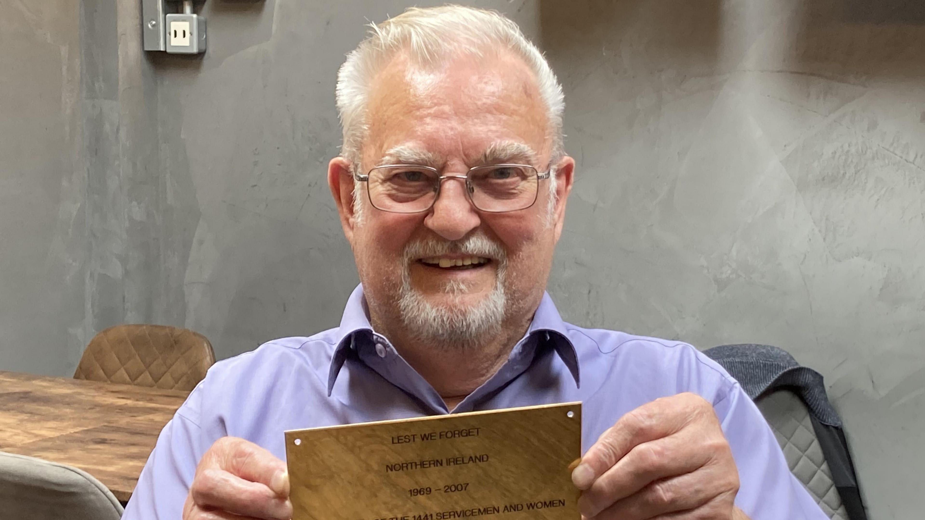 Roger Moseley holding up one of the plaques that will be on the memorial. He is wearing a blue shirt and smiling at the camera