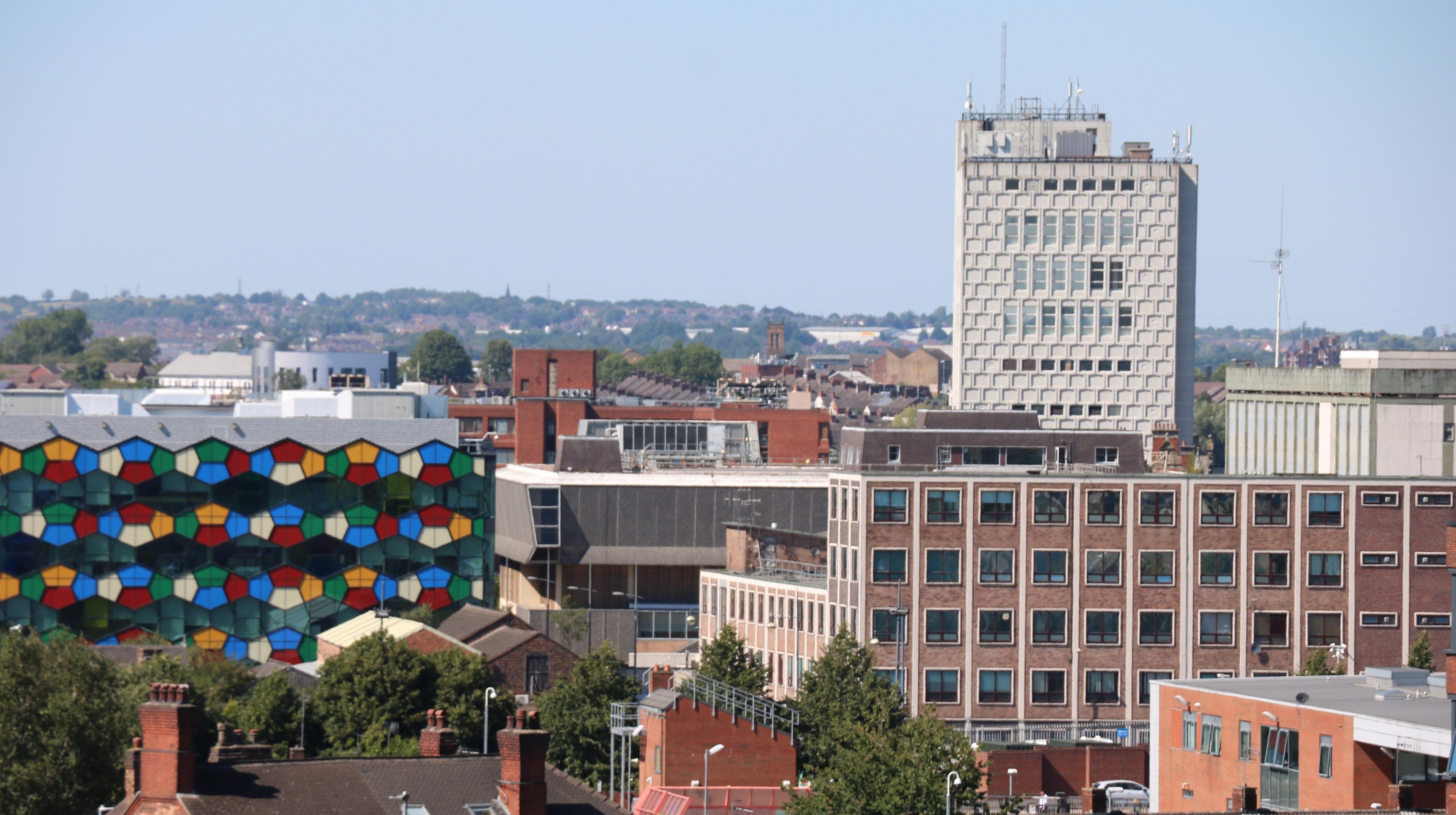 From left, a multicoloured glass building appears among a series of others, a tall white building in the background rises up above 