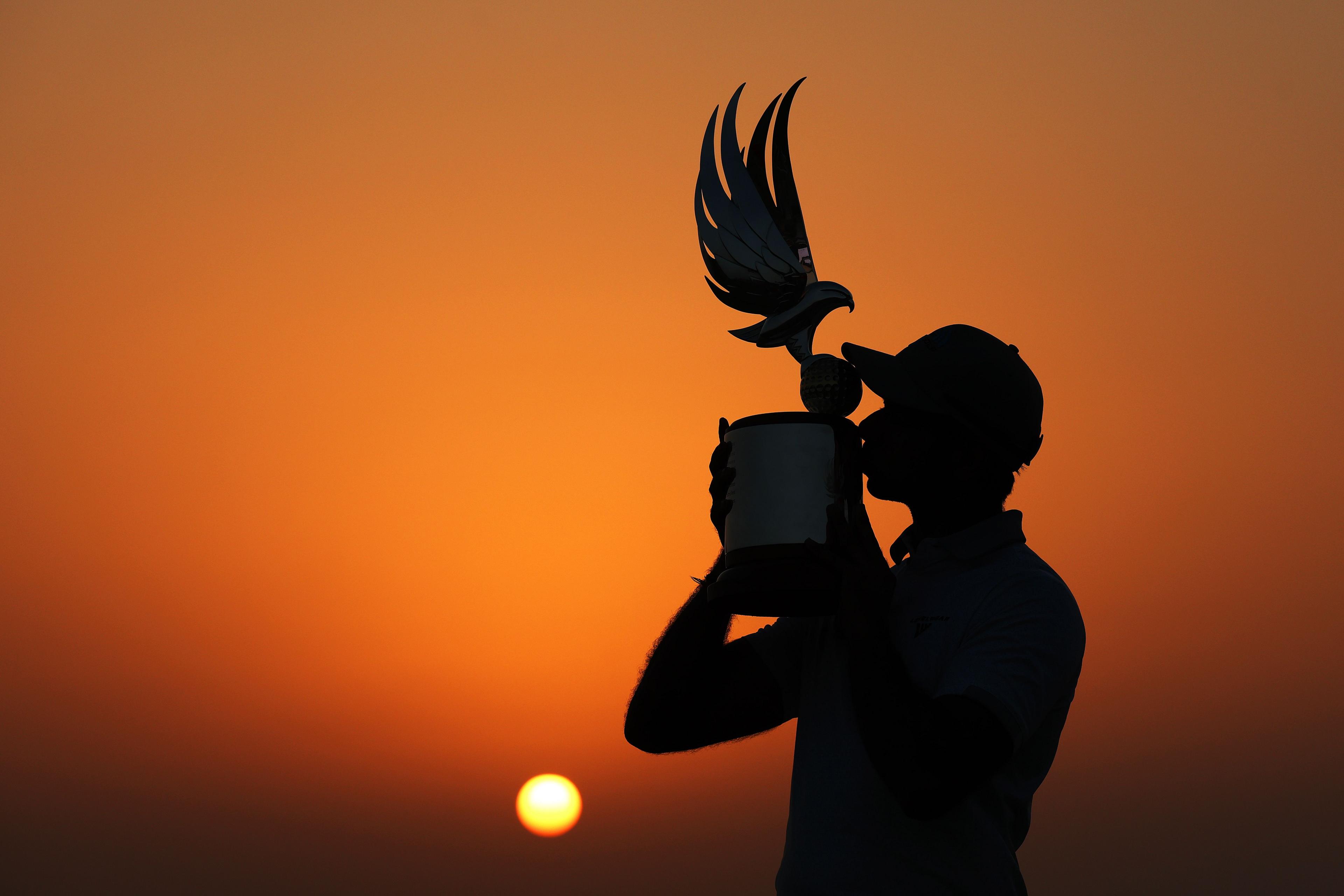 Silhouette of a person holding and kissing a trophy with a winged design against a vivid orange sunset, with the sun low on the horizon.