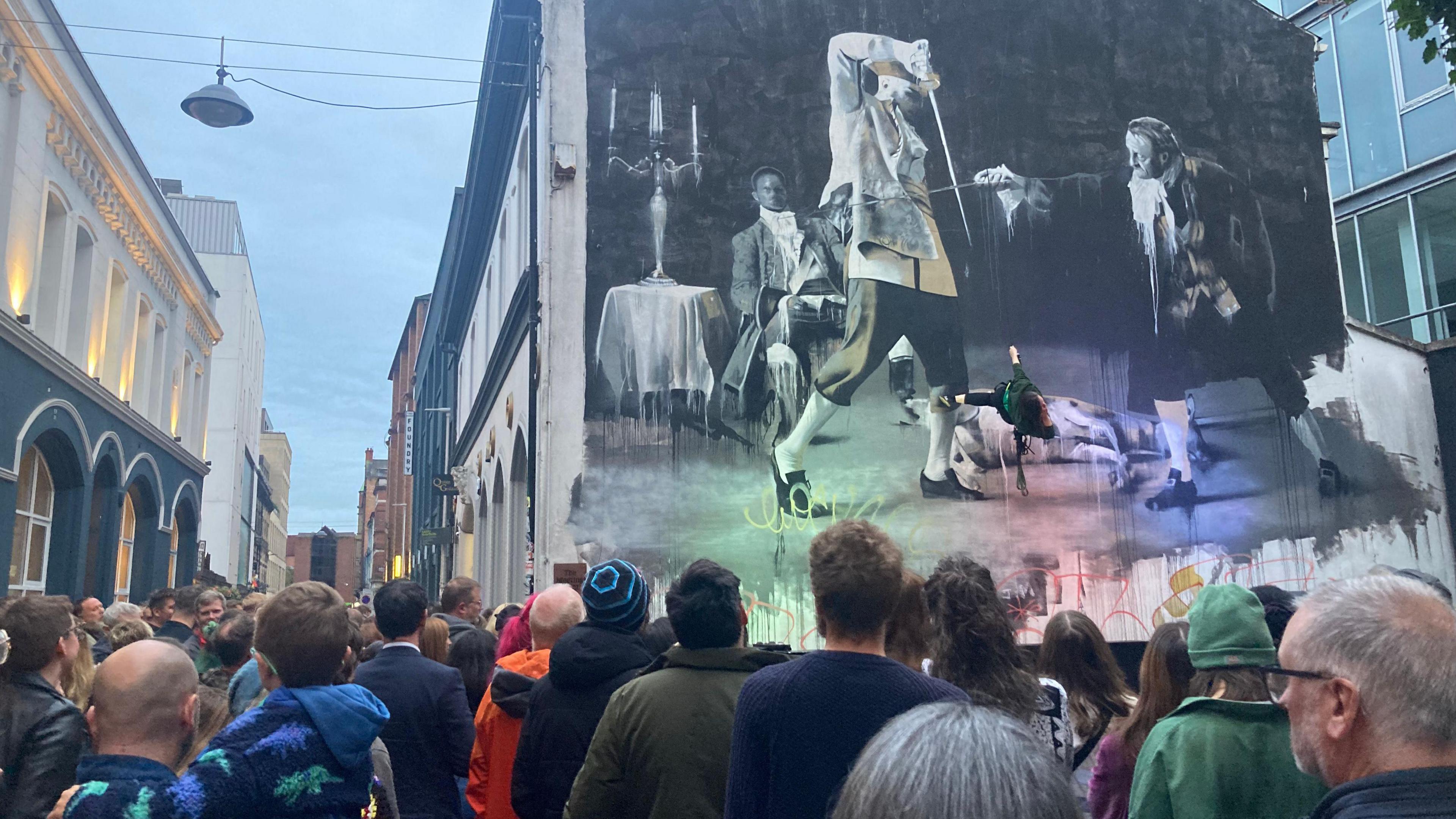 A number of people are seen near a large gable wall. The artist is suspended from a wire. 