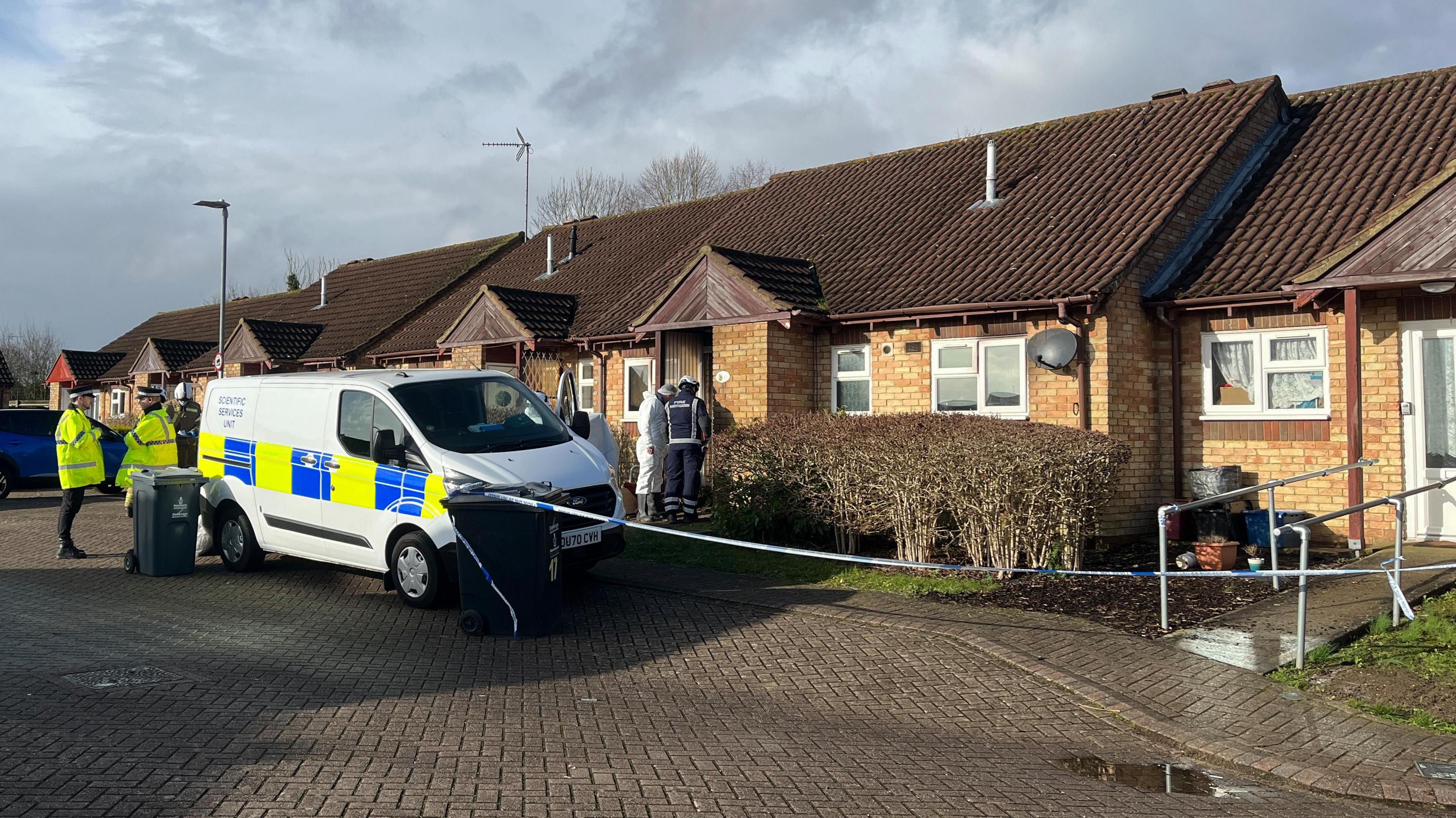 A row of bungalows are cordoned off by police tape. There are police officers and fire crews visible. There are forensic teams dressed in white stood nearby too. There is a van parked outside that says "scientific services unit" on the side.