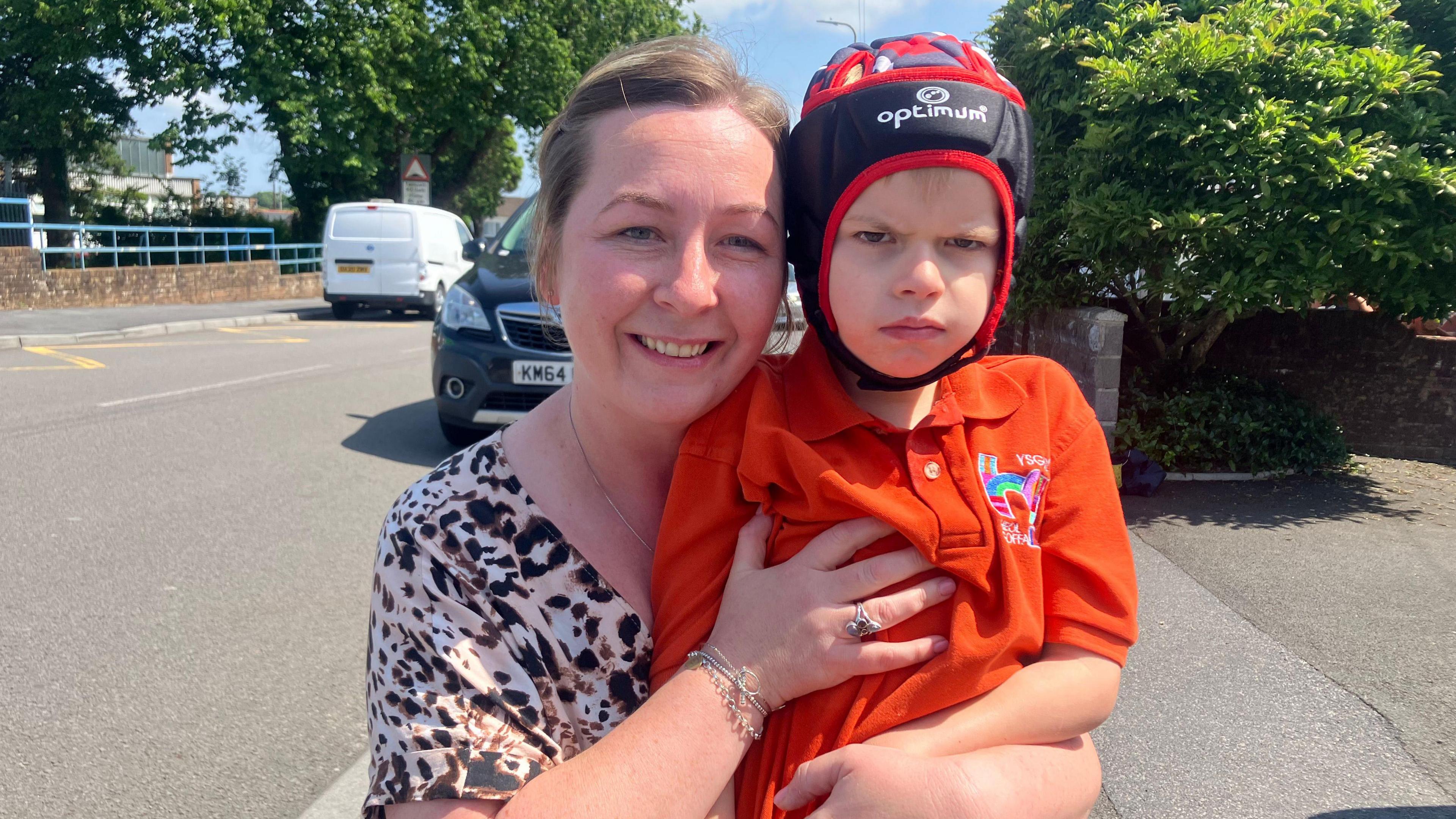 Lana Dakin (left) holding her son Jac, who attends Ysgol Heol Goffa. Jac is wearing a bright orange polo shirt and helmet, Lana has her blonde hair tied back and is wearing a leopard print shirt.