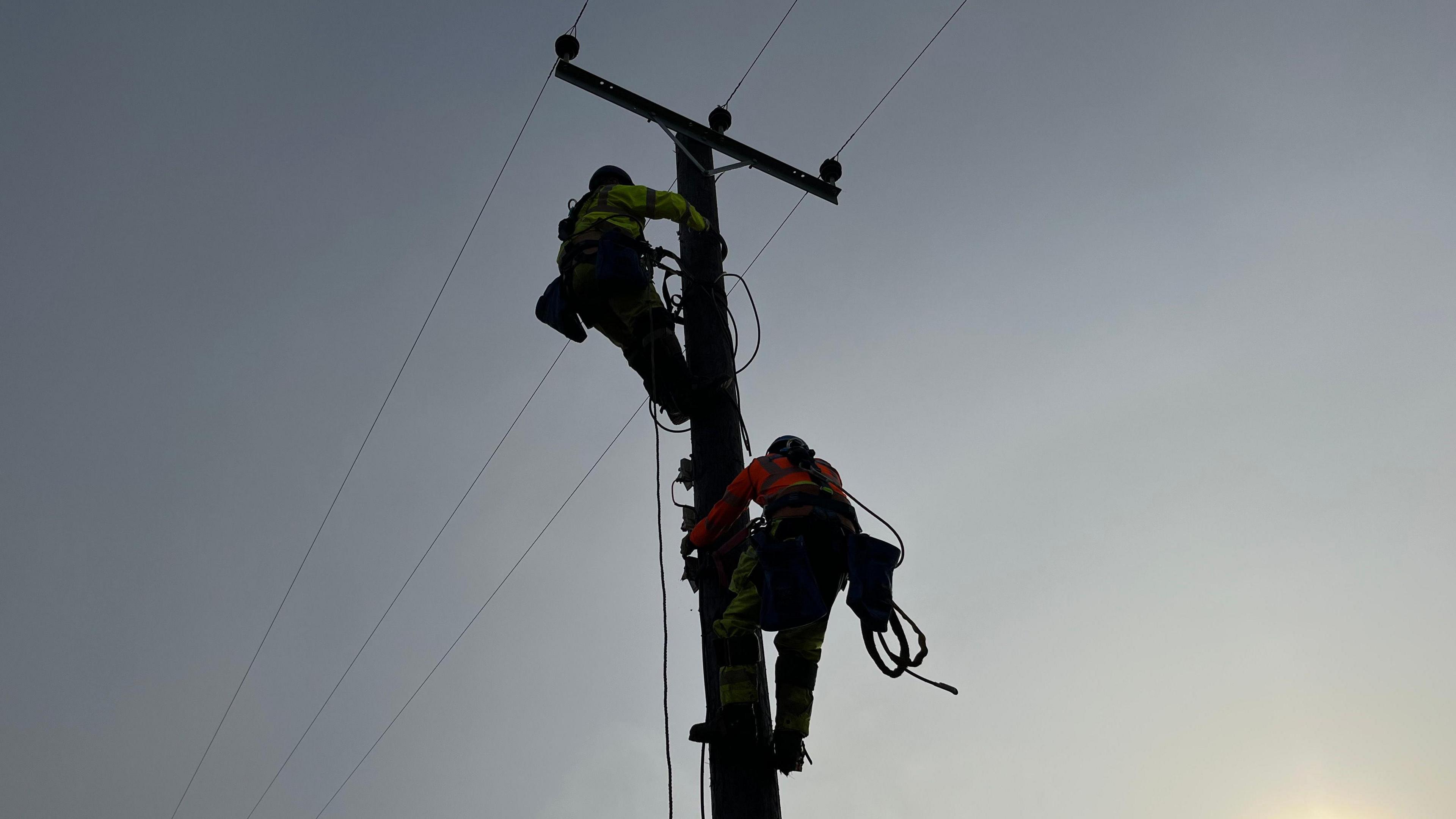 A worker in a yellow high vis jacket above another worker in an orange high vis jacket up an electricty pole with a light blue sky. 