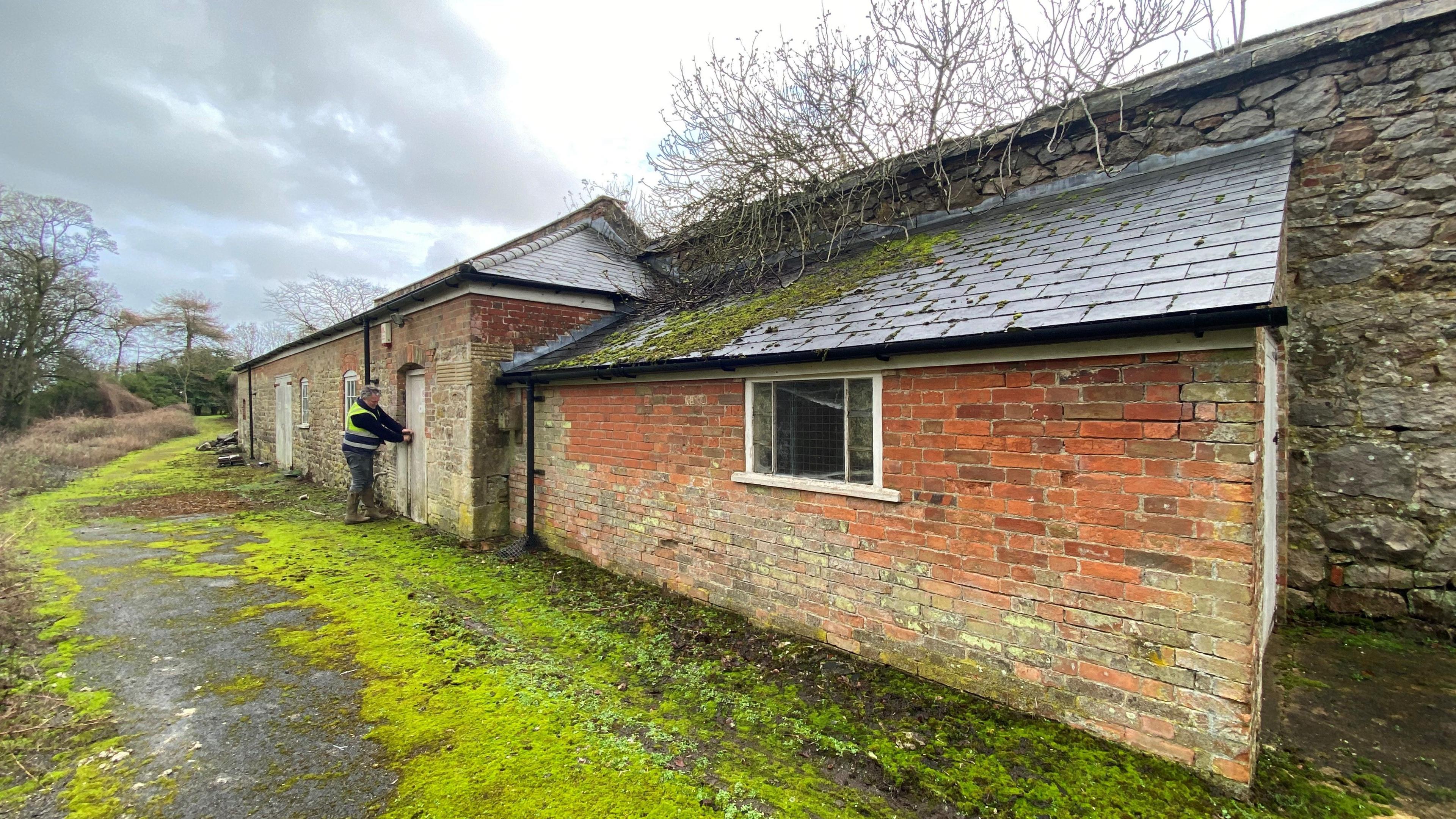 The disused bothy on the perimeter of the walled garden. It looks very old and abandoned, with bricks that are coming away from the mortar, mouldy windows, moss and dead branches on the roof, and algae growing everywhere. There is a main building on the left, and a one-storey extension on the right. The bricks are cracked and faded, and there is a man in a hi-vis vest pulling the wooden door closed. 