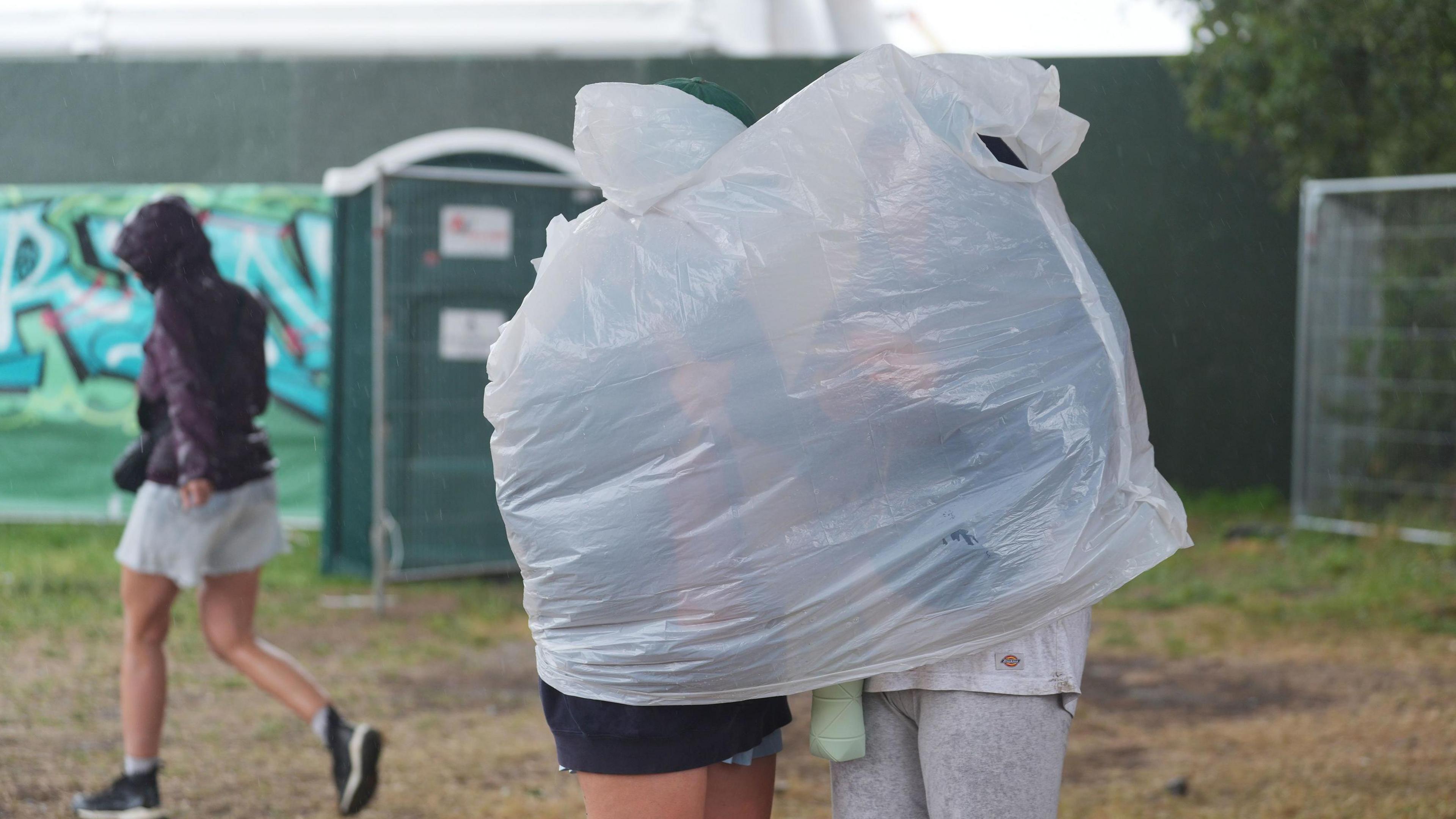 Two people standing near a green portaloo at the Glastonbury Festival, with a white plastic poncho stretched over both of them.