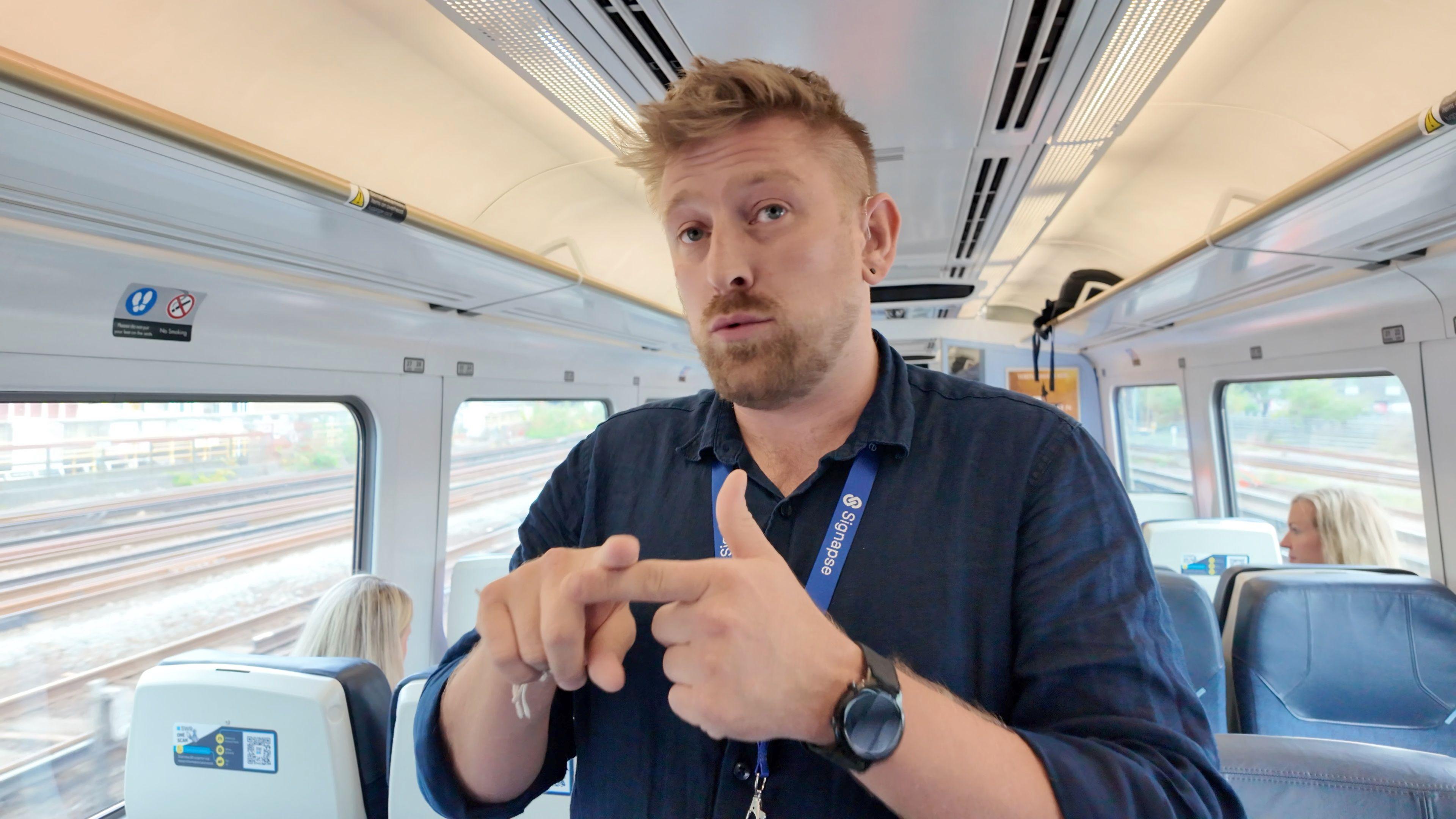 Marcus Oaten is standing in the aisle of a train, he has a couple of passengers in the background behind him and is giving an interview using British Sign Language. Marcus has short hair and a beard, he wears a navy blue collared linen shirt, a black watch on one wrist and a stack of bracelets on the other