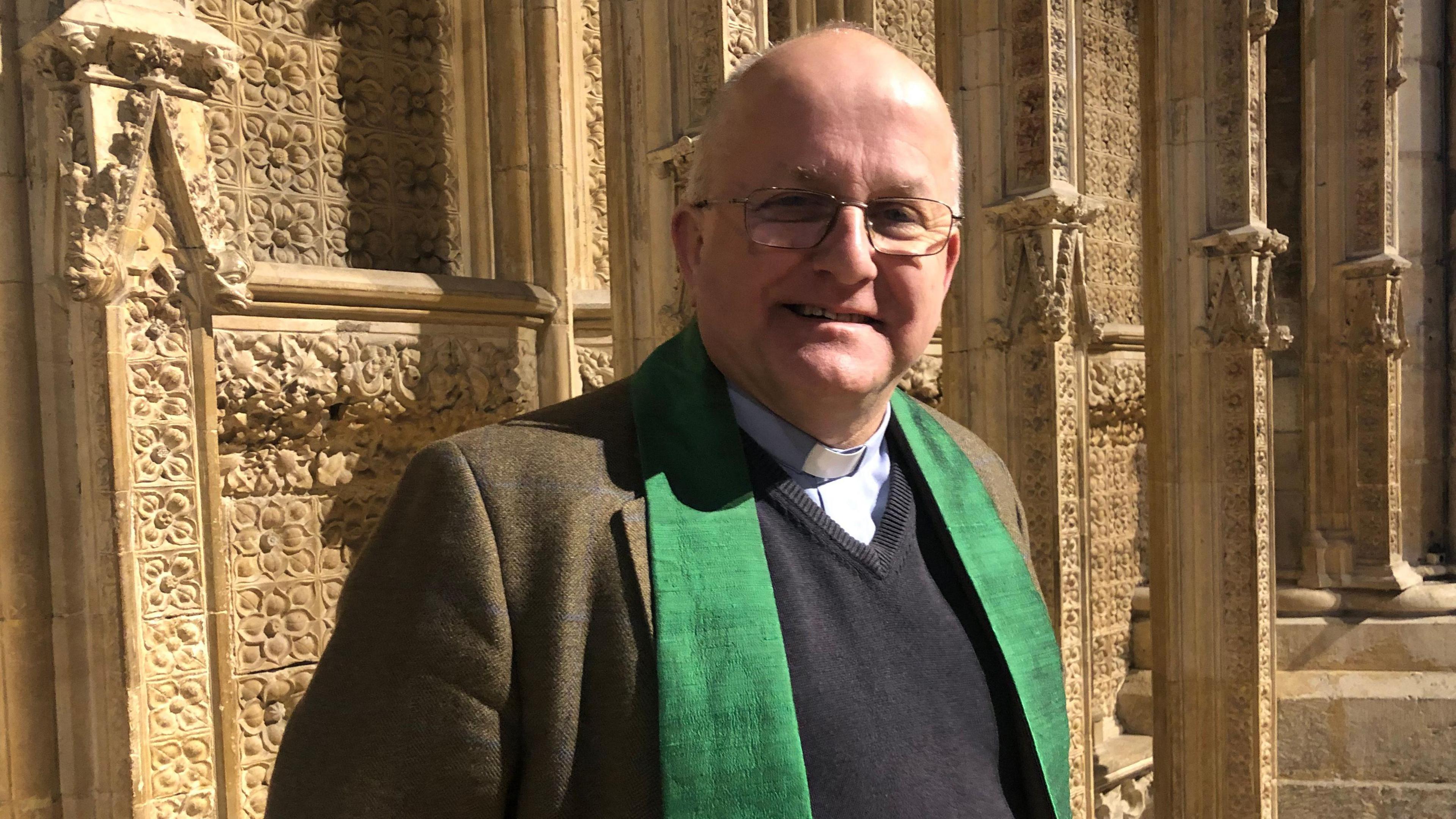 A man stands in front of stonework at Lincoln Cathedral: wearing glasses, a clerical collar, a tweed jacket, and a green stole.