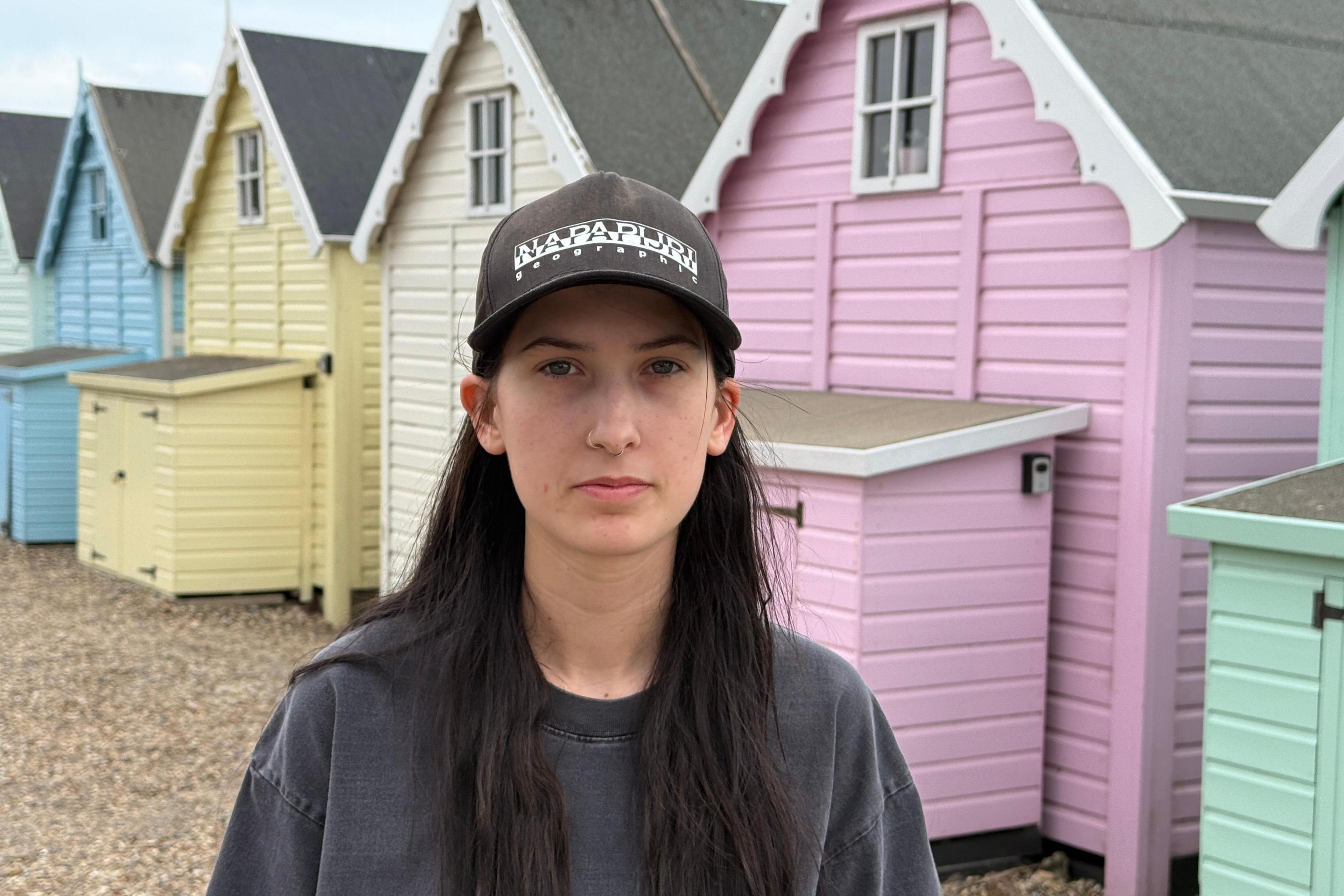 Leonie Powling stands on a stony beach, in front of multicoloured beach huts. She wears a dark-coloured jumper and black baseball cap.