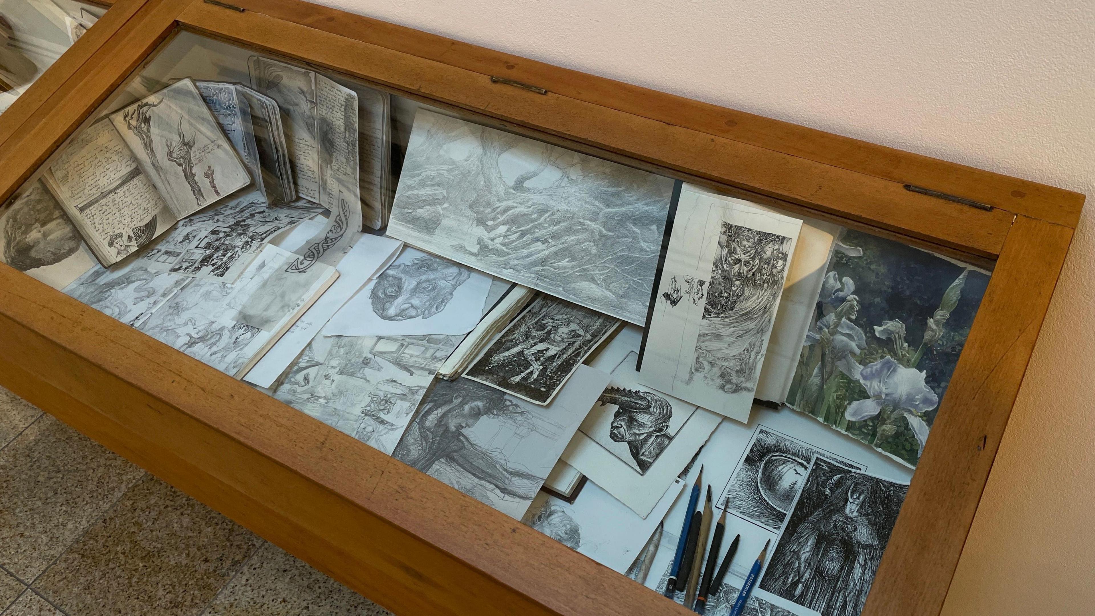 A display case full of sketches. Some show woodland or mythical creatures. One sketchbook is open showing the first illustrations for Gandalf's staff.