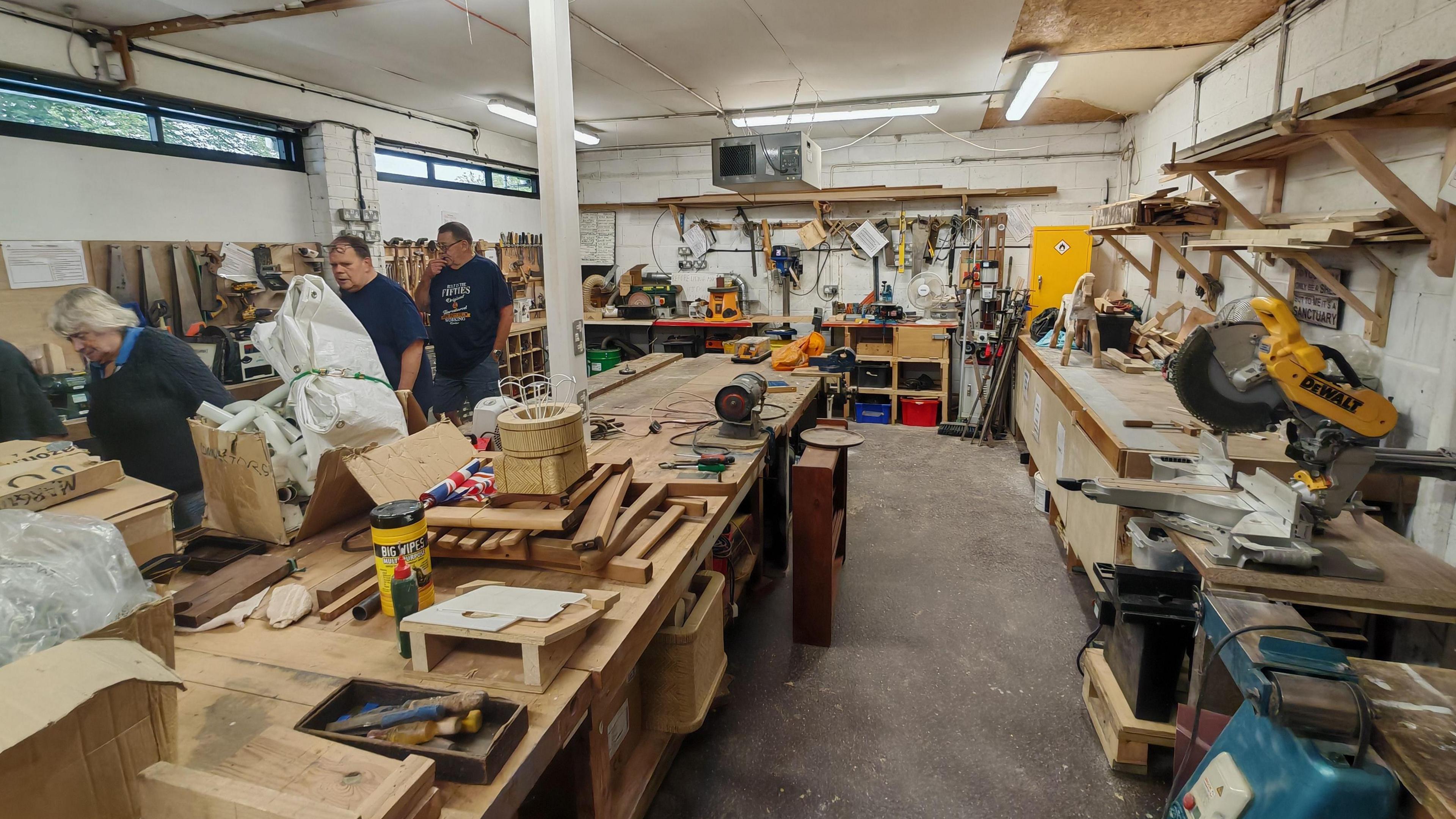 The interior of the Spelthorne Shed, with several volunteers standing at workbenches, of which there are many, covered with tools.