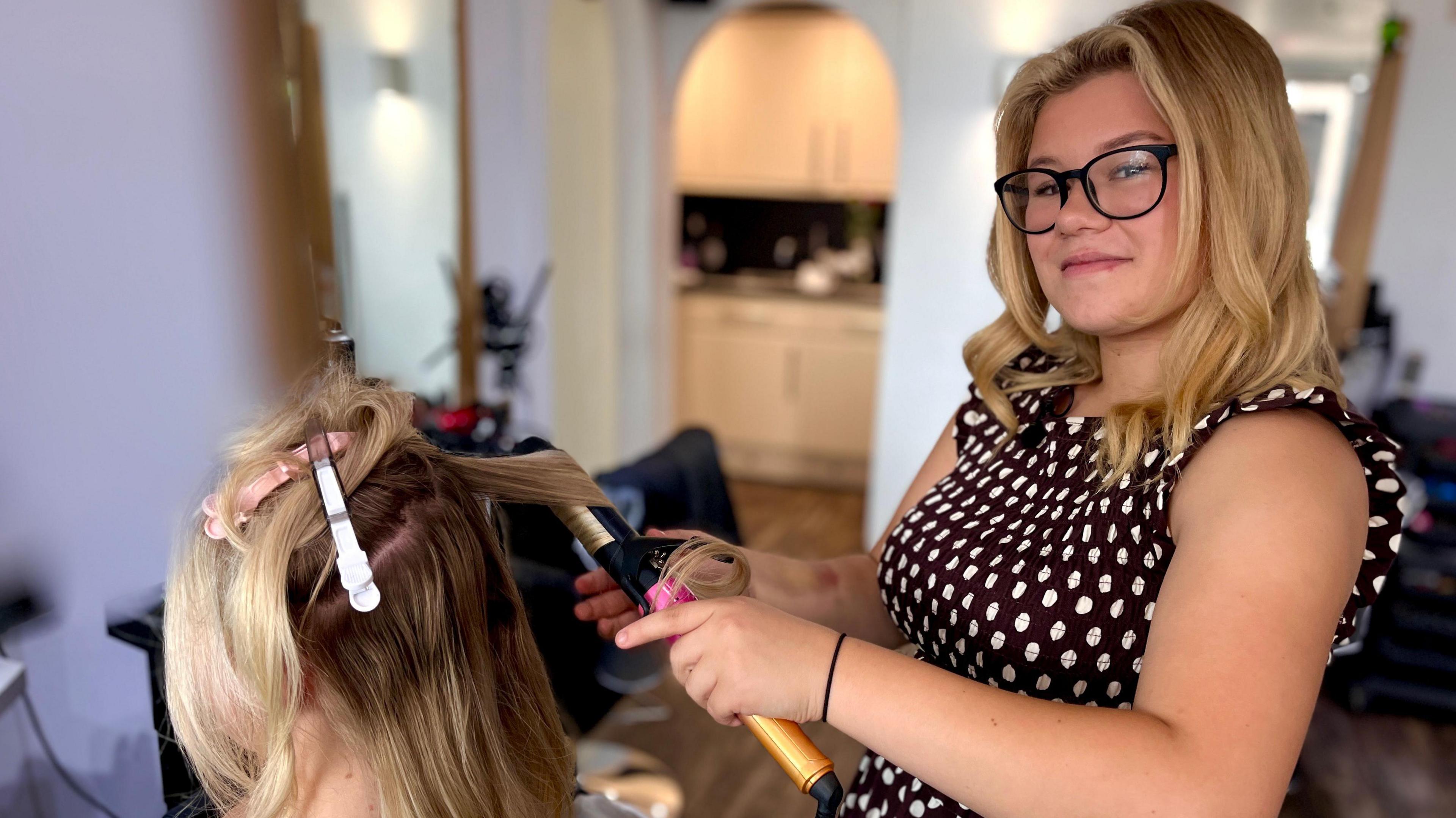 Jessica Flaxman stands in a hair salon, wearing a brown polka-dot dress and black-rimmed glasses, with her long blonde hair visible. She is using curling tongs on a client seated in front of her. The client, who also has blonde hair, has several styling clips sectioning her hair.