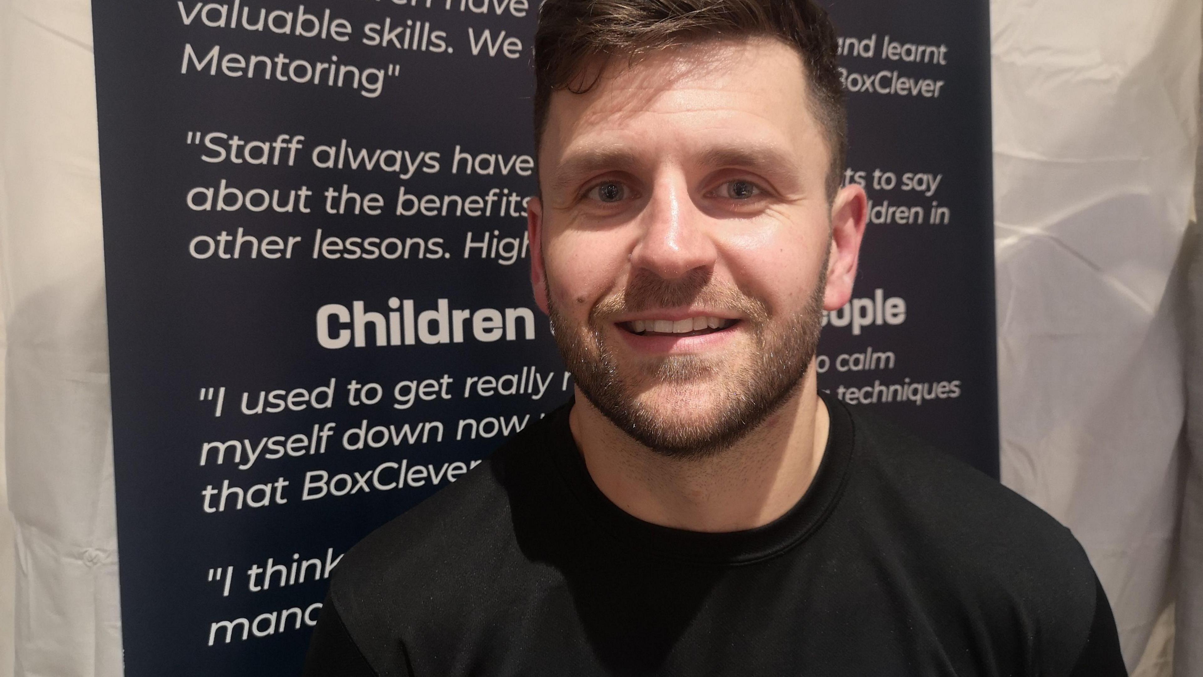 A man wearing a black t-shirt smiles into the camera. Behind him is a poster with quotes.