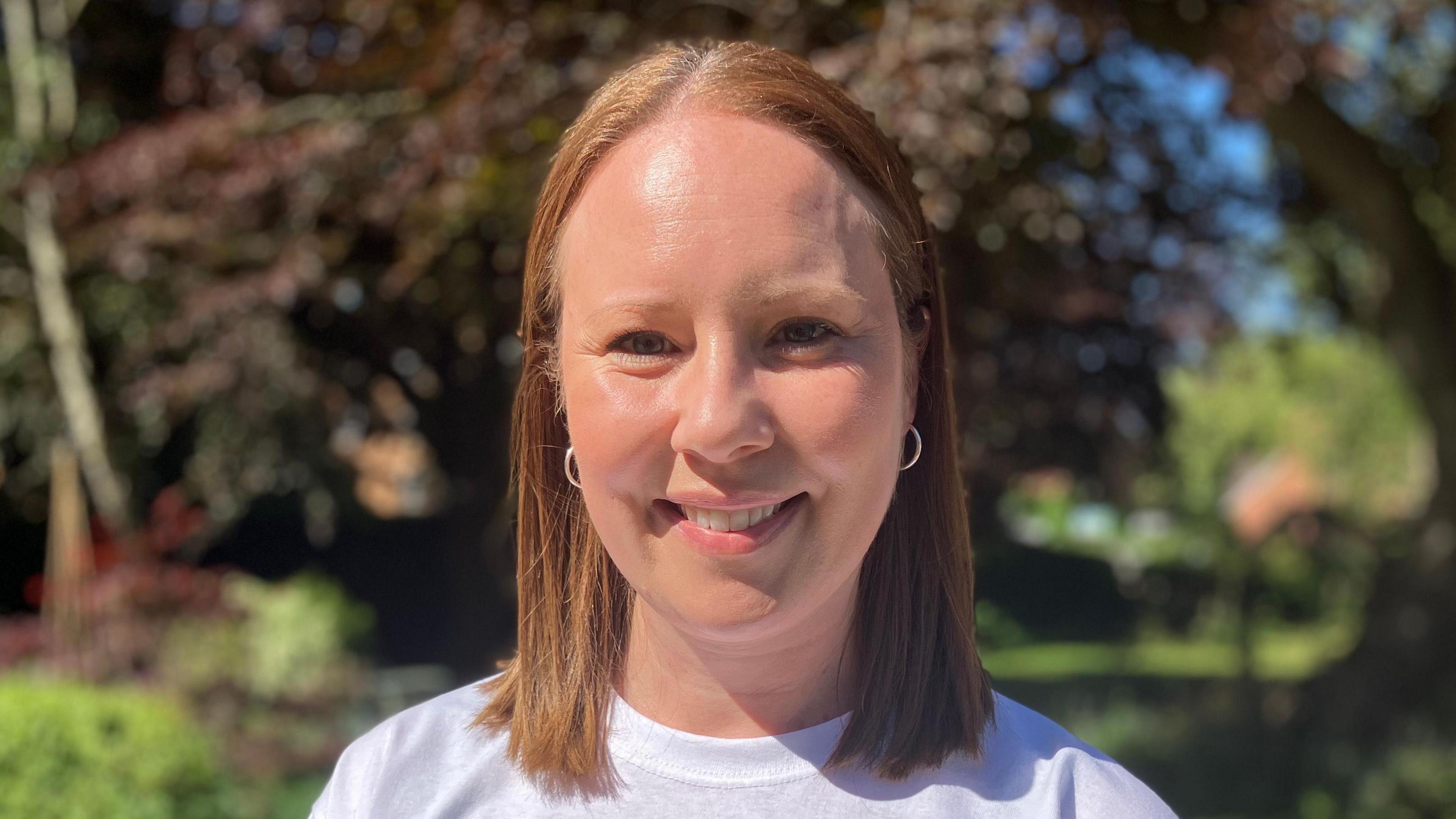 Sam Perkins's wife Emma Perkins, a white woman with short brown hair, wearing a white t-shirt and small hoop earrings.