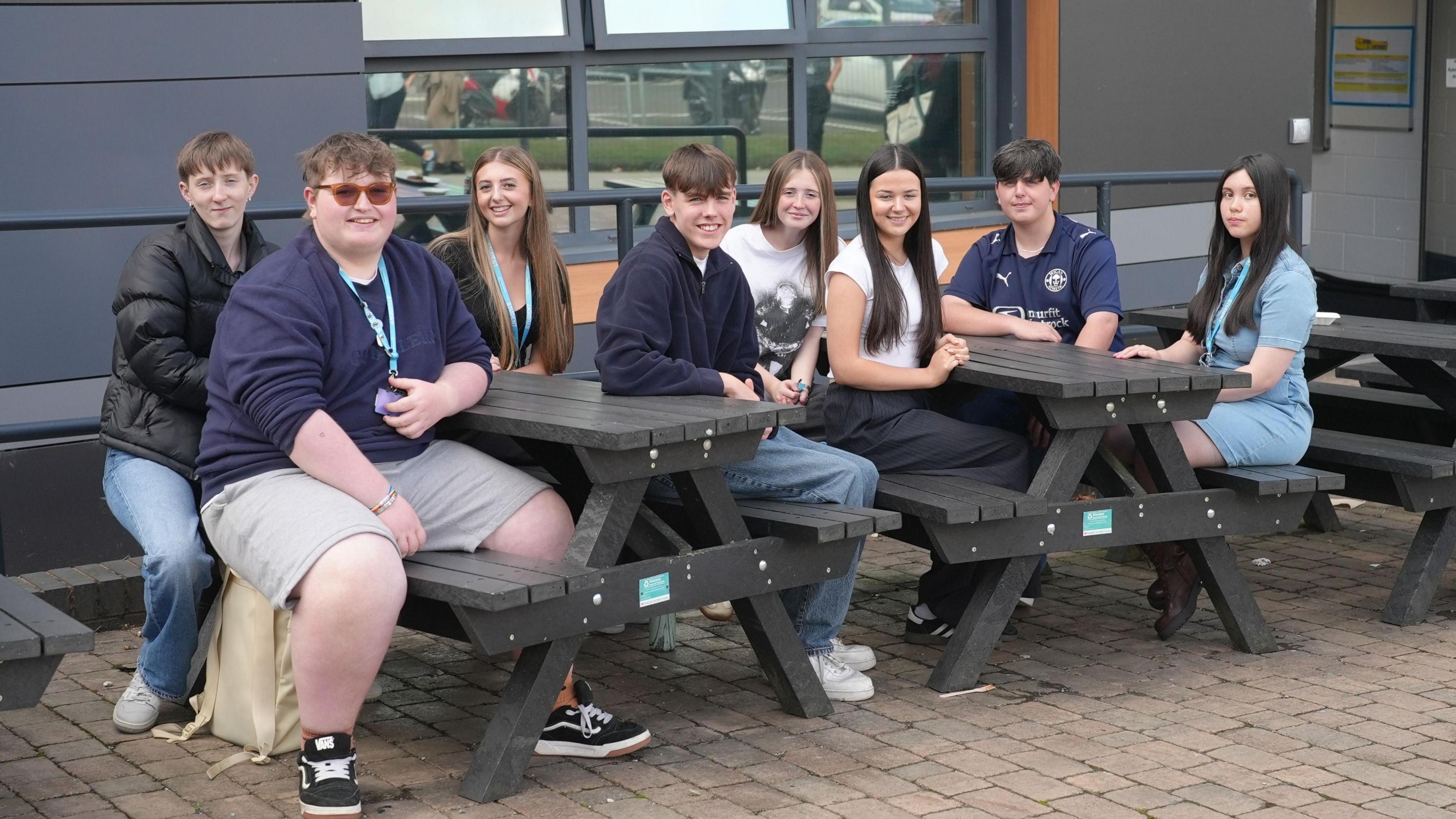 A group of male and female students, all aged 17, sit together, smiling, at two picnic tables in front of their college building