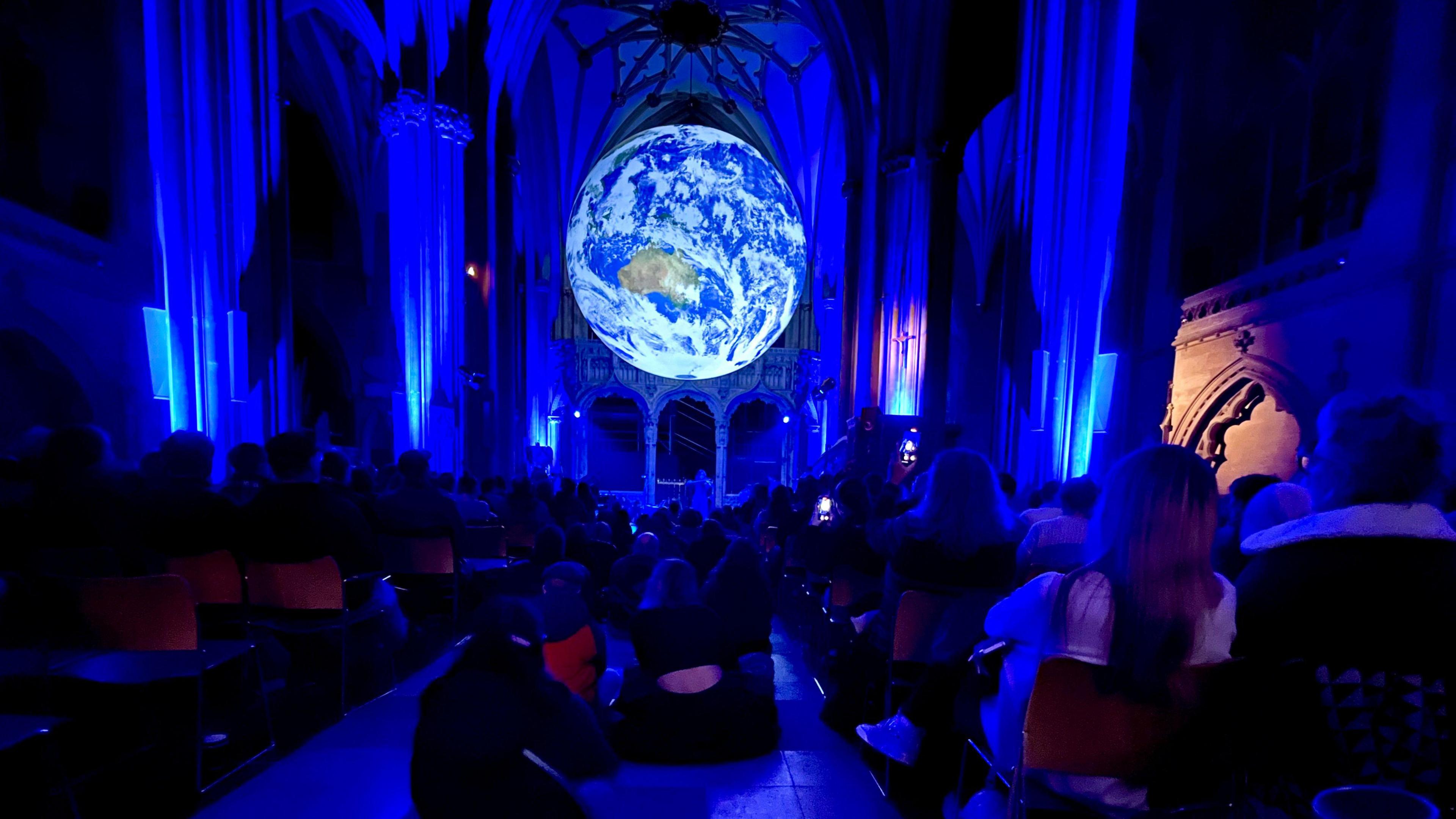 People watch a musical display under the Luke Jerram artwork Gaia inside Bristol Cathedral. The internal columns of the cathedral are illuminated by blue light and Gaia, a to-scale model of the earth, is illuminated