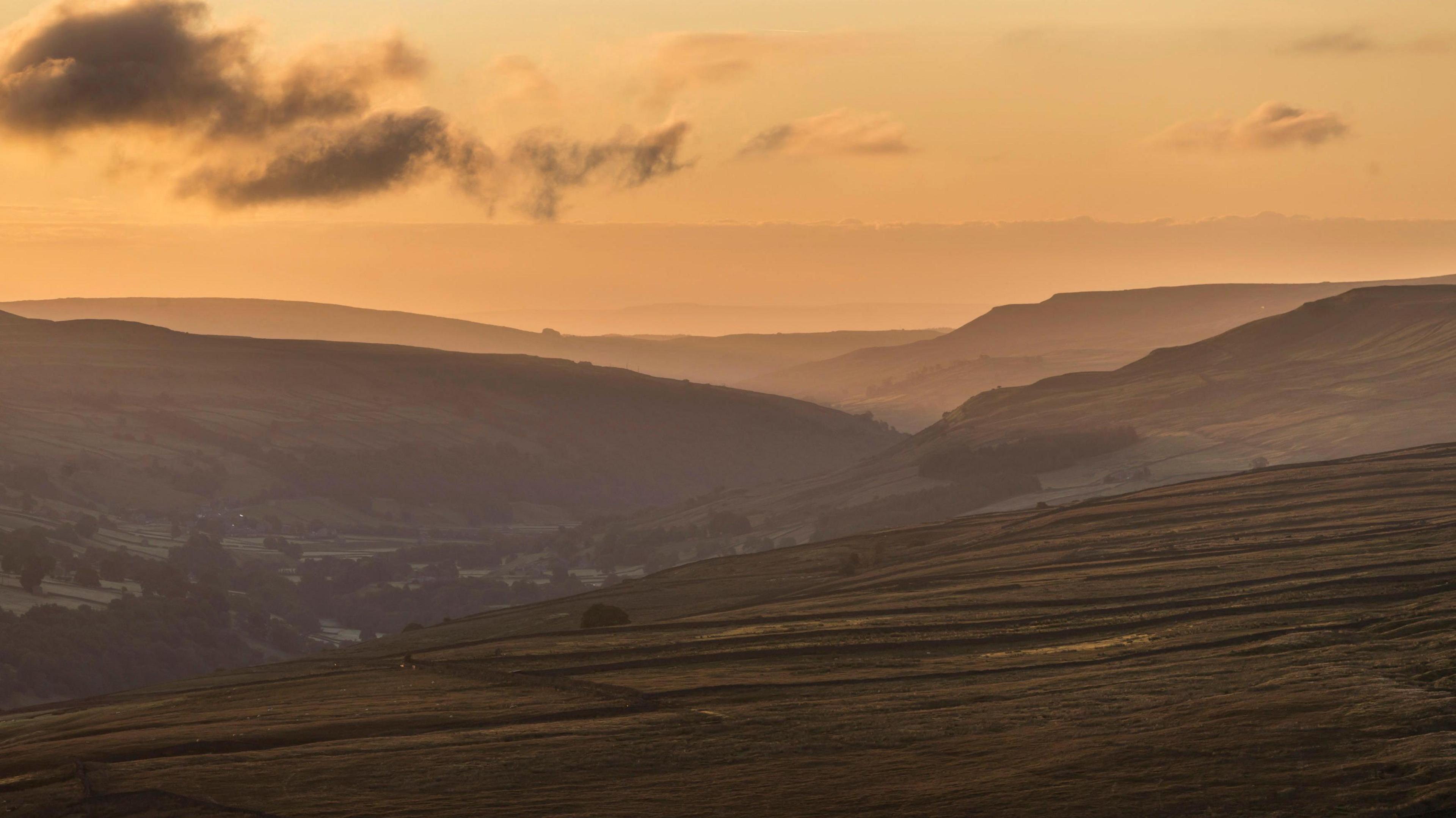 A view over hills and fields, with a yellow sky and slight mist in the distance