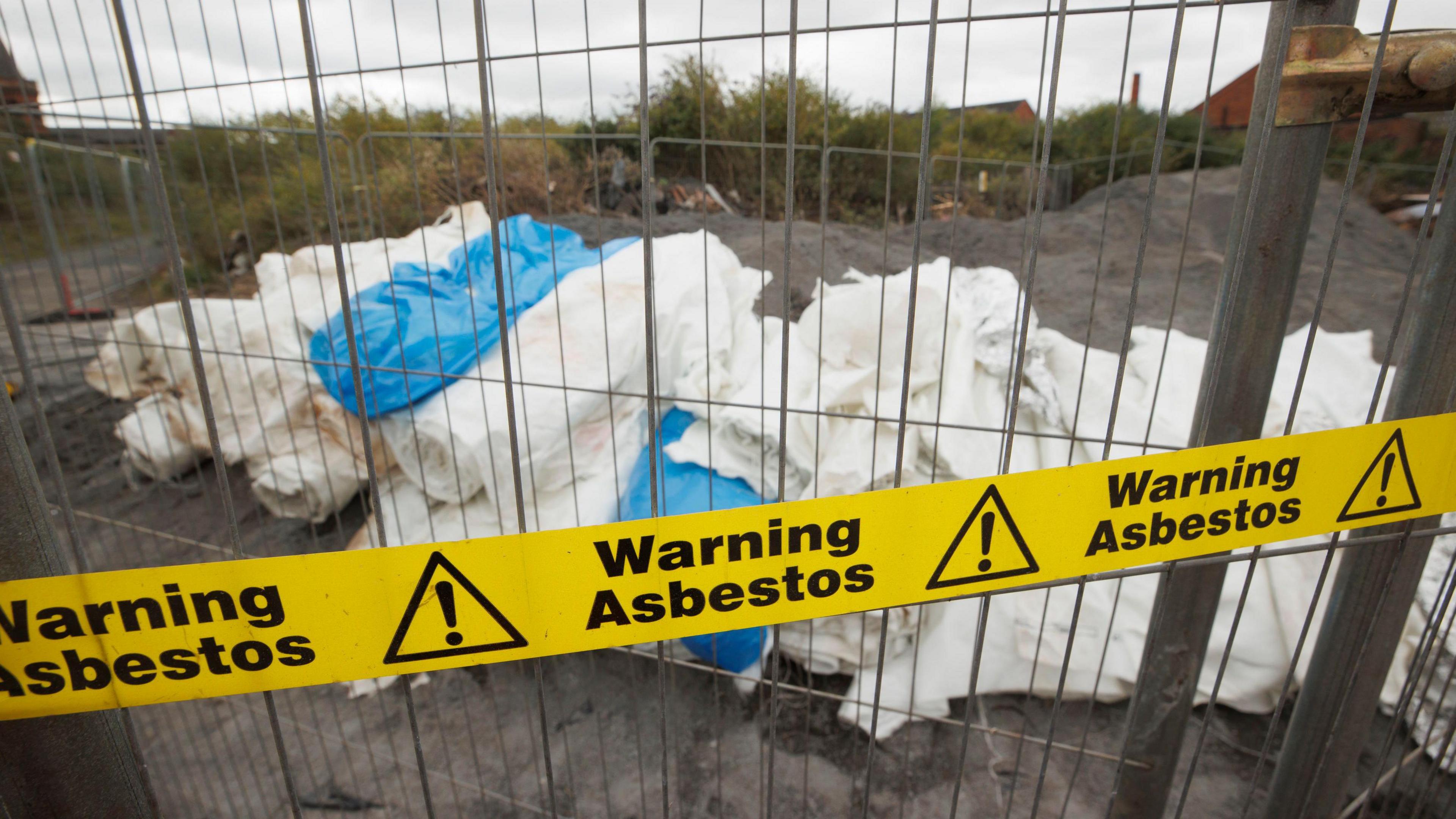 A pile of plastic sheets rolled up on the ground, with a metal fence and yellow tape reading "warning asbestos" in front of it. 