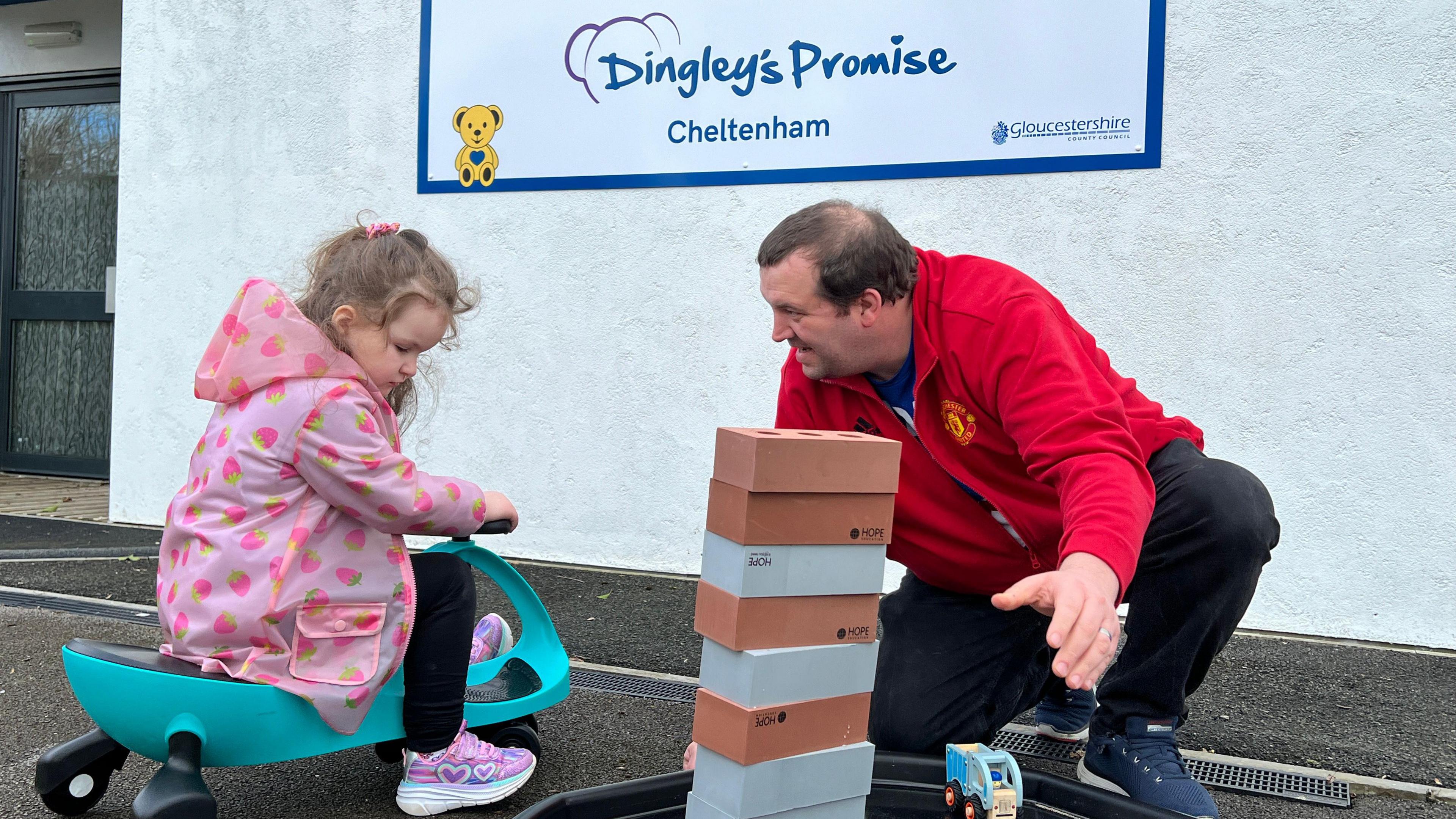 A girl is wearing a pink spotty coat and trainers with hearts on them, is playing on a green plastic trike in the playground of a children's centre, whilst her dad in a red jumper smiles at her next to a pile of toy bricks stacked up on top of each other.