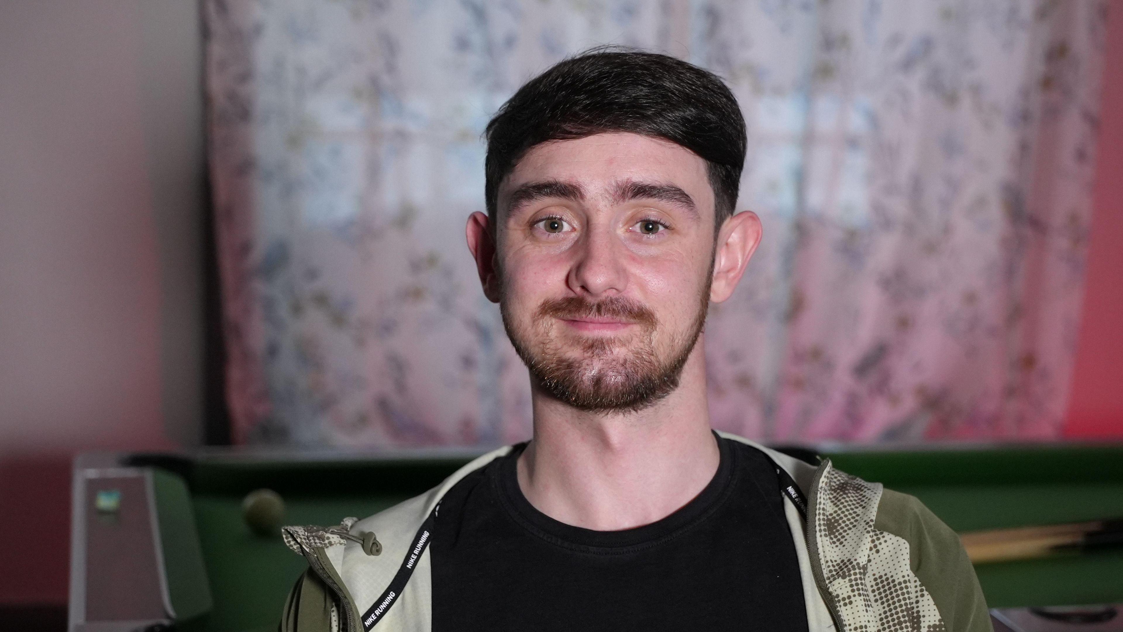 A head and shoulders picture of a man in his 20s looking straight into the camera and smiling slightly. He has short dark brown hair, dark prominent eyebrows and brown eyes. He has a short brown beard and moustache and is wearing a black t-shirt with an olive-green, patterned tracksuit-style jacket open over the top.  A pool table is visible behind him, and some white patterned curtains are drawn across the window behind that. 