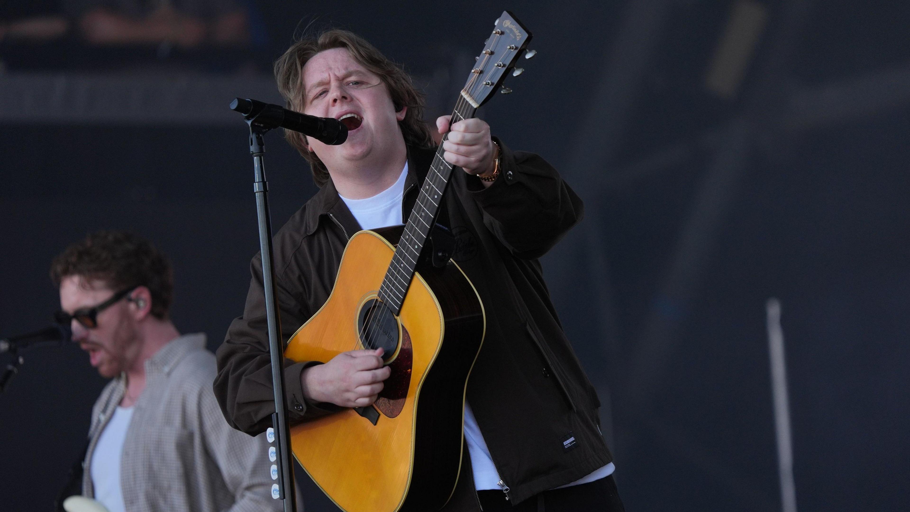 Lewis Capaldi, who has long brown hair and is wearing a brown jacket over a white t-shirt, sings into a microphone on stage while playing a light brown acoustic guitar.