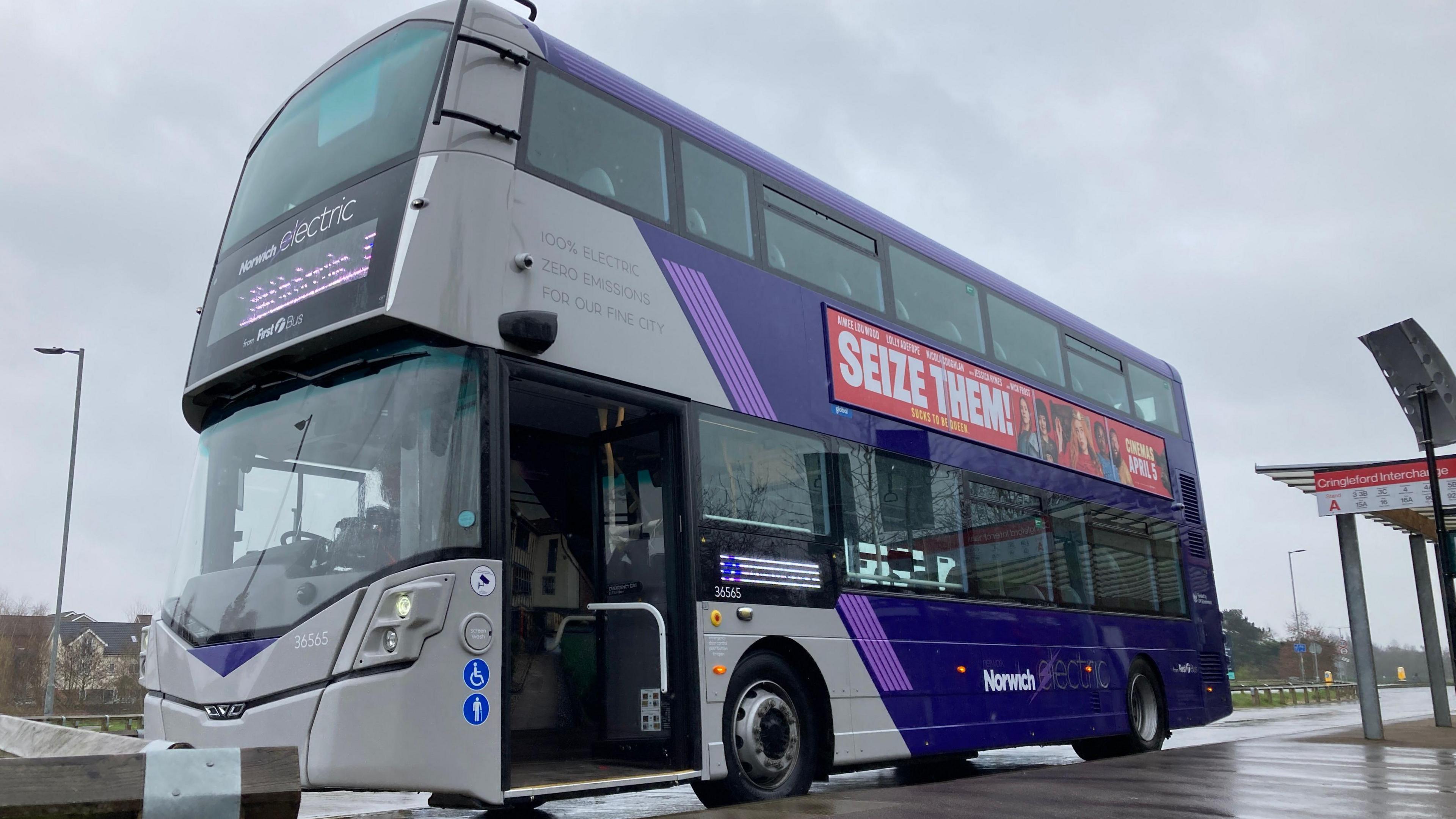 A purple and grey electric double-decker bus is parked at a bus stop on a rainy day. The bus door is open and a promotional sign on the side of the bus reads 'seize them'. The bus is a First bus. 