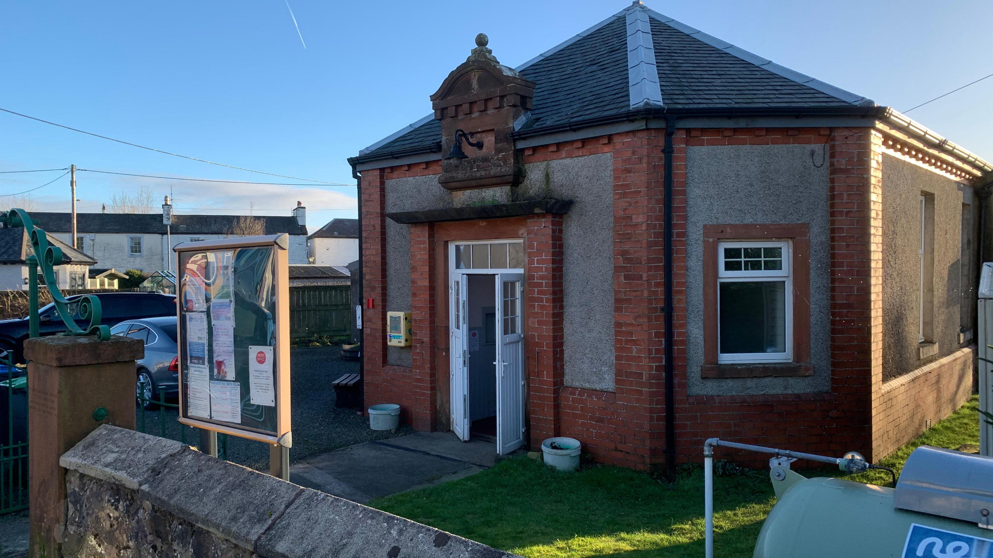 An old brick building with a notice board in front of it surrounded by a low sandstone wall