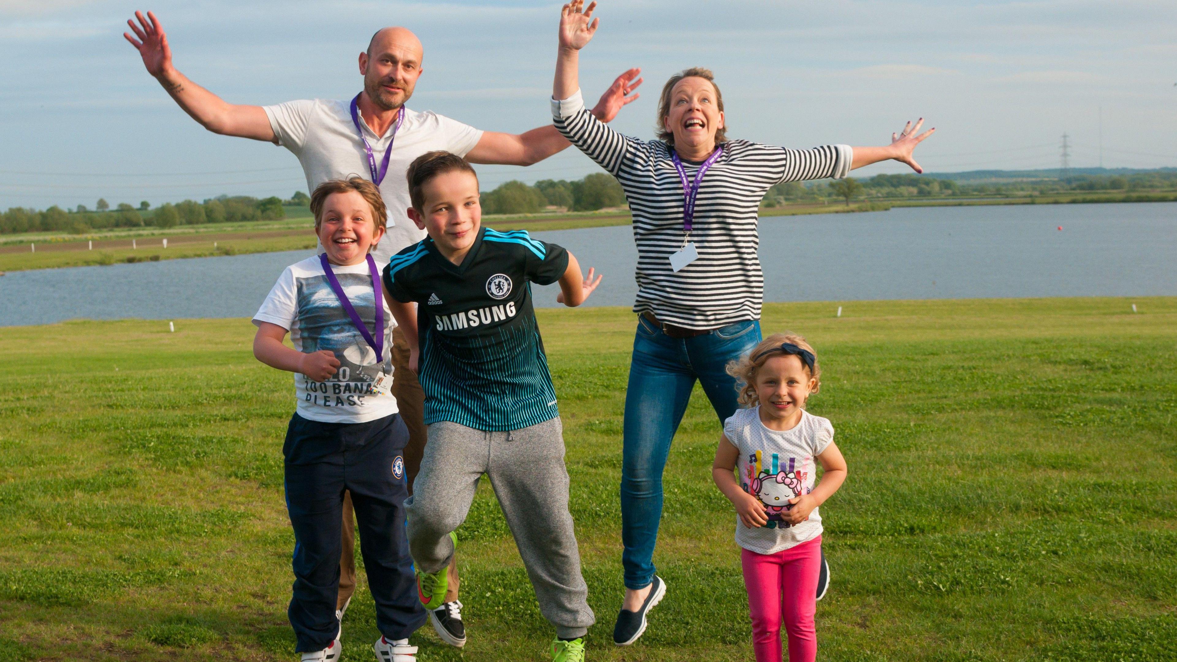 Two adults and three children are jumping and have their arms in the air on a field  with a lake in the background.