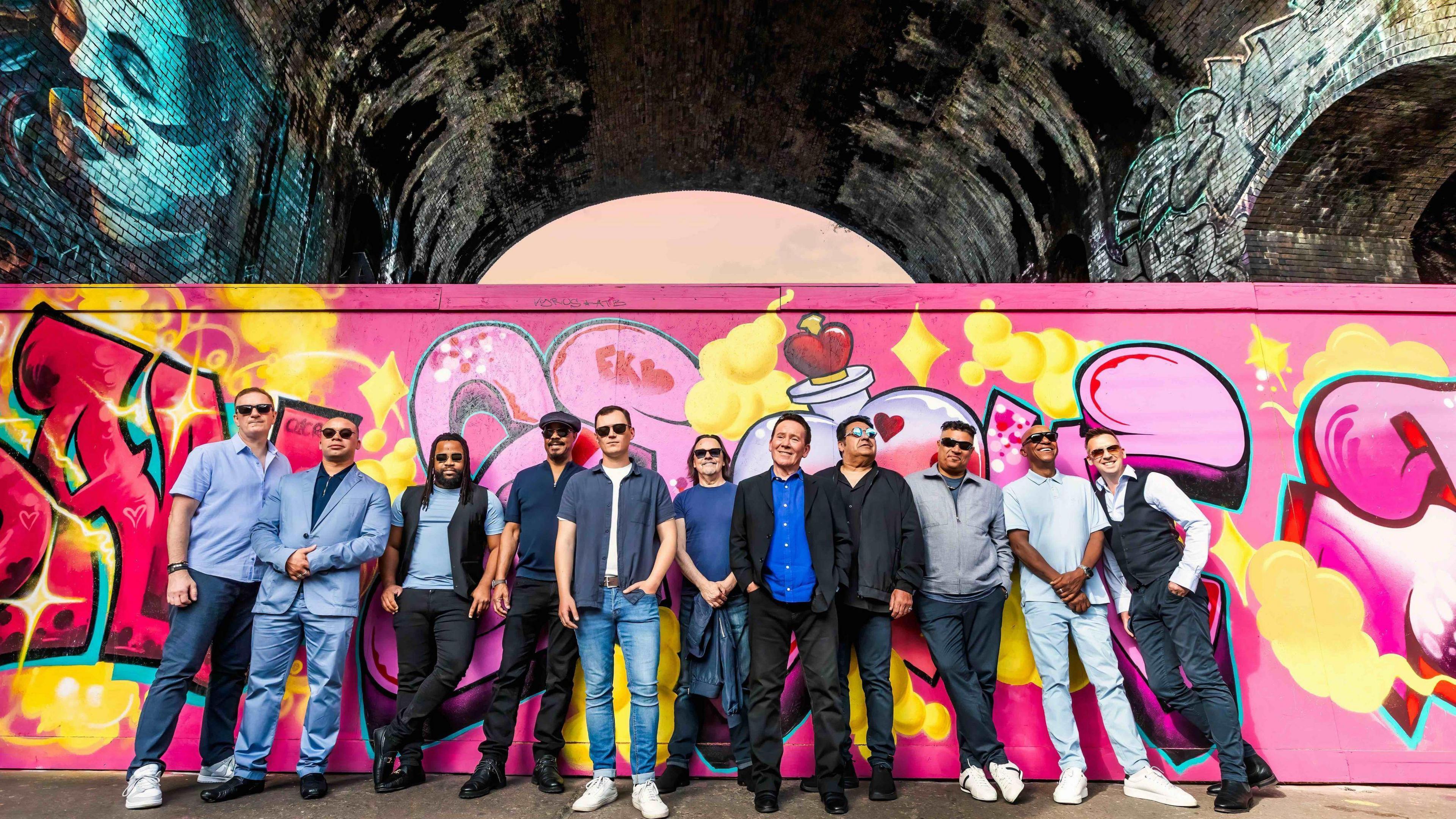 The band, standing together in front of a largely pink graffiti wall