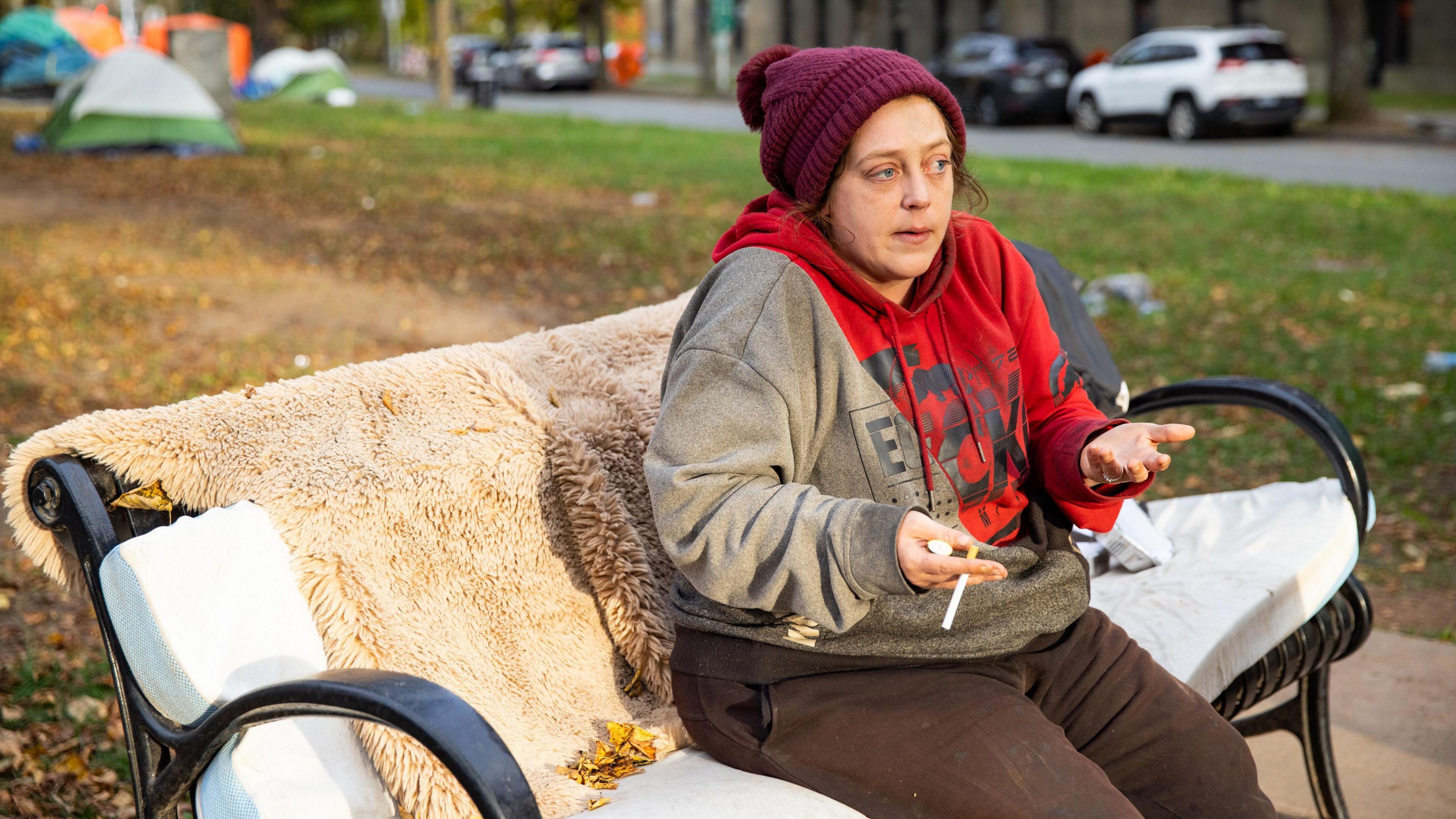 Samantha Nickerson sits on a park bench in downtown Halifax. She wears a burgundy toque, a grey and red hoodie and is speaking animatedly with a cigarette in hand 