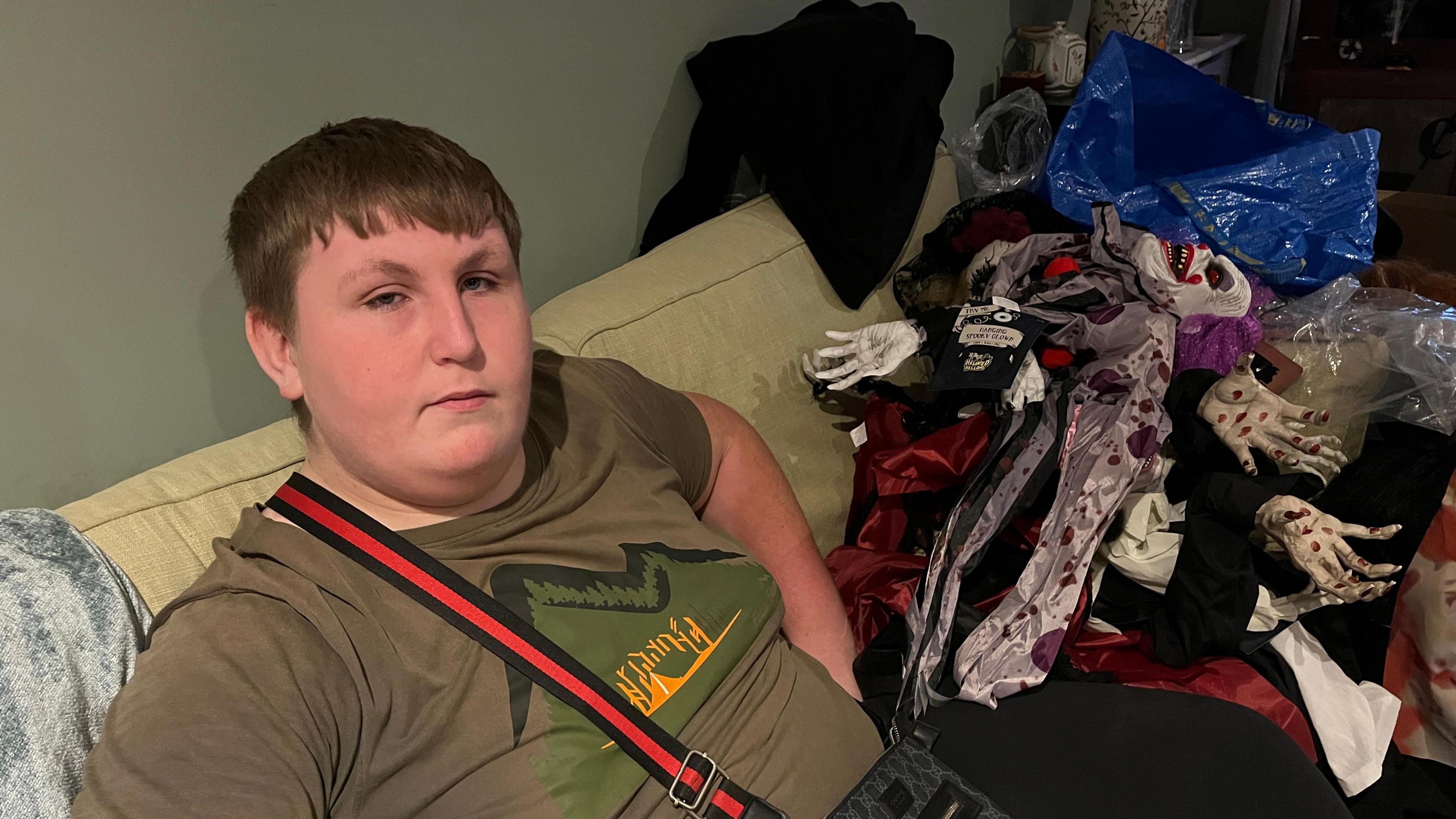 Adam Wright sits on a sofa inside his house. He has short brown hair and wears a green T-shirt with a bag resting over his torso. Next to him on the sofa are Halloween decorations in the form of clowns. 