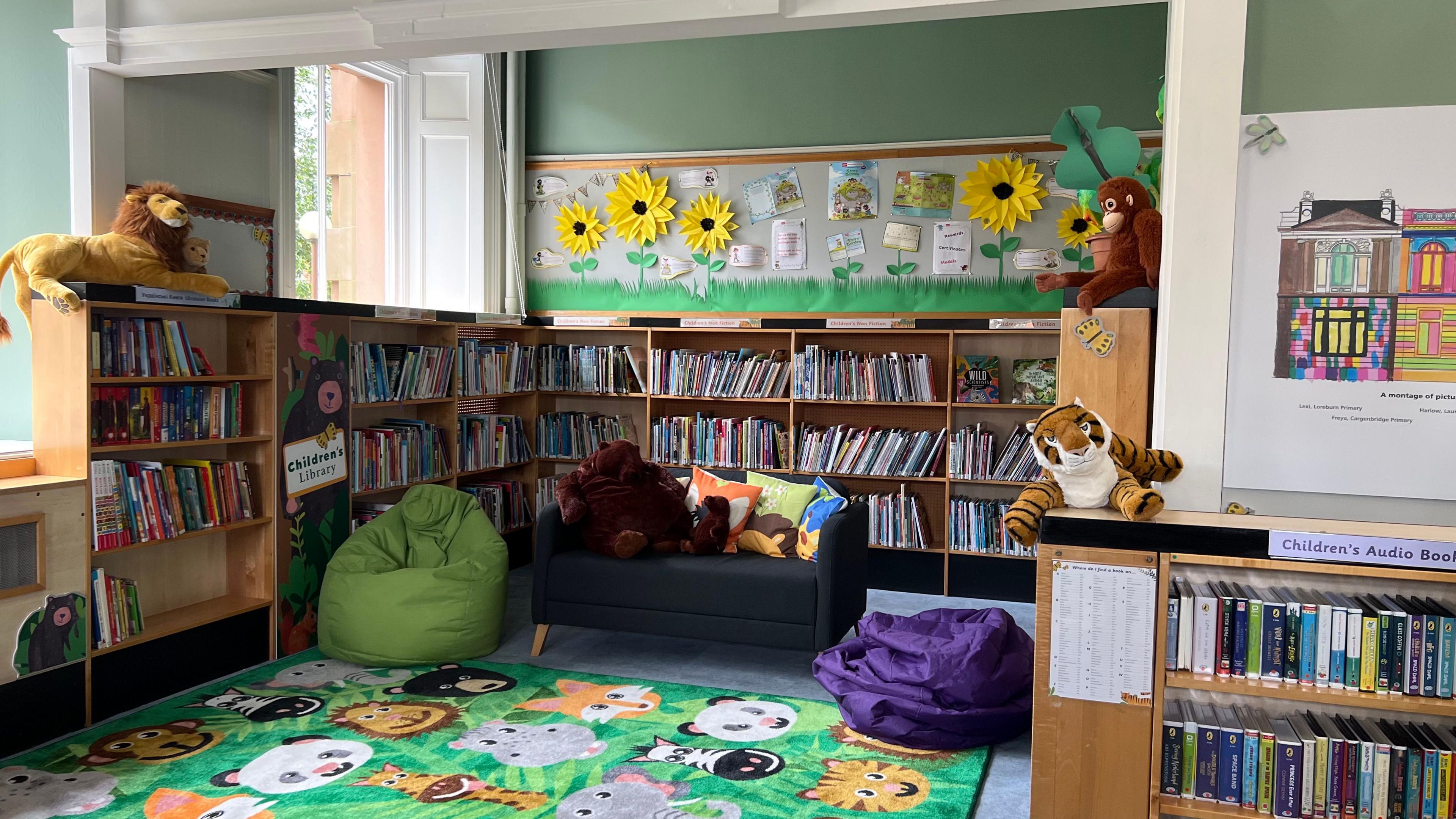 A nook surrounded with book shelves. In the centre a grey couch with two stuffed bears, it is flanked by two bean bags - one green, one purple. The floor has a green rug with different safari animals on top.