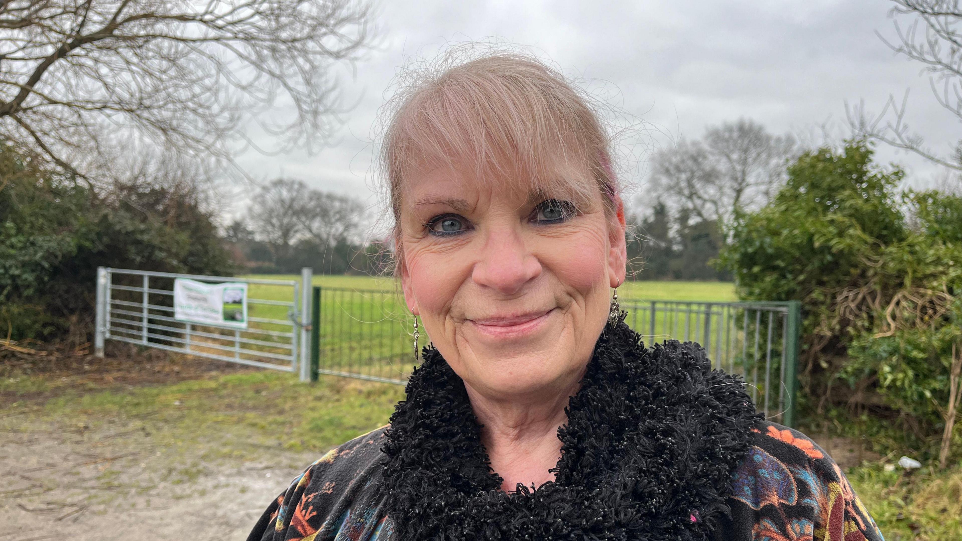 Teri is looking into the camera and smiling. She's wearing a black coat with an orange and blue floral pattern on it, and a black woolly scarf. Behind her you can see the silver and green fencing blocking access to the Glebe, and beyond that  the Glebe itself, a large green field with trees and shrubbery around its edges. 