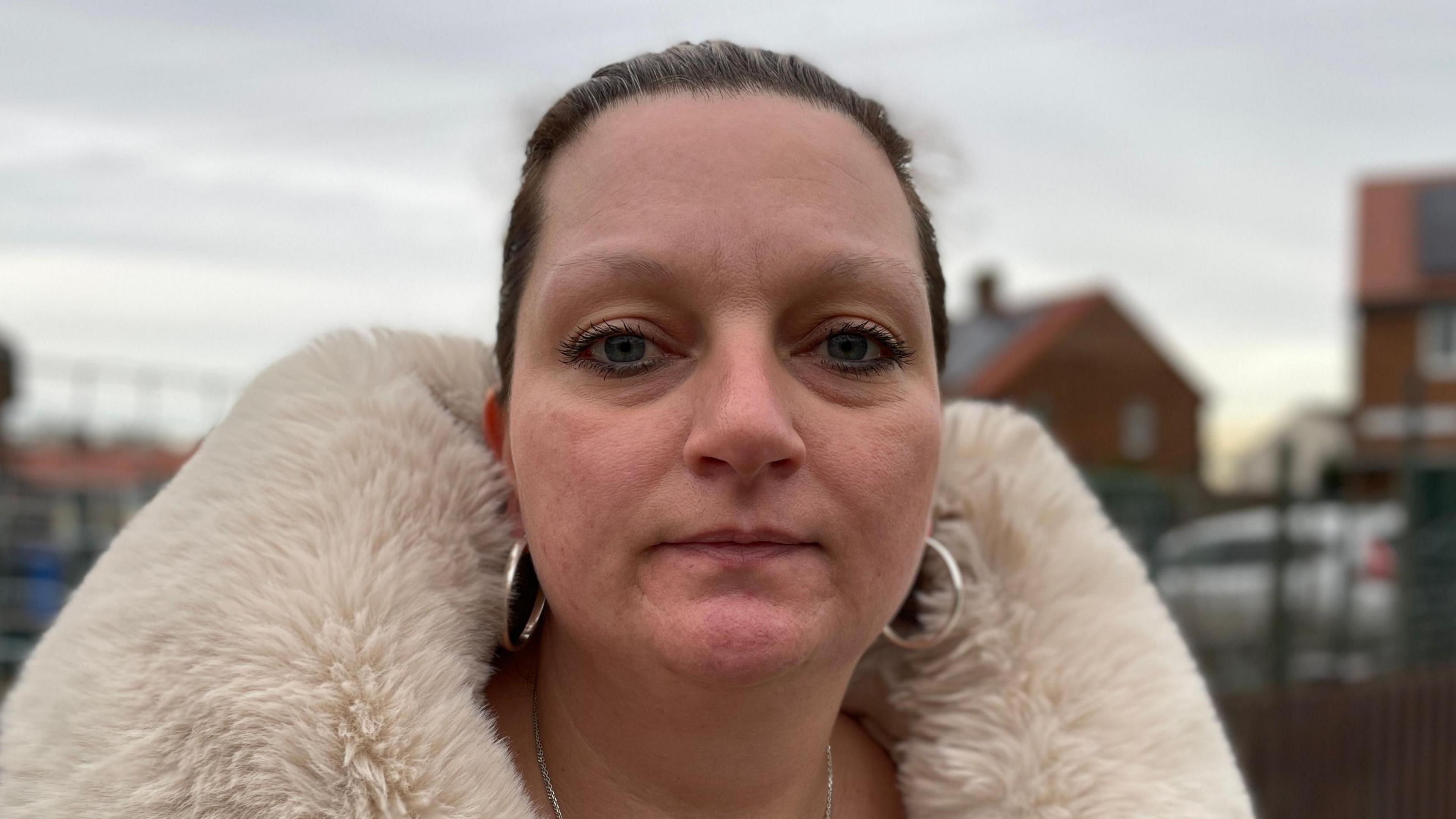 Tracey Englefield looks at the camera. She has dark hair that is tied behind her head. She wears silver hoop earrings and has a cream-coloured fur coat on.