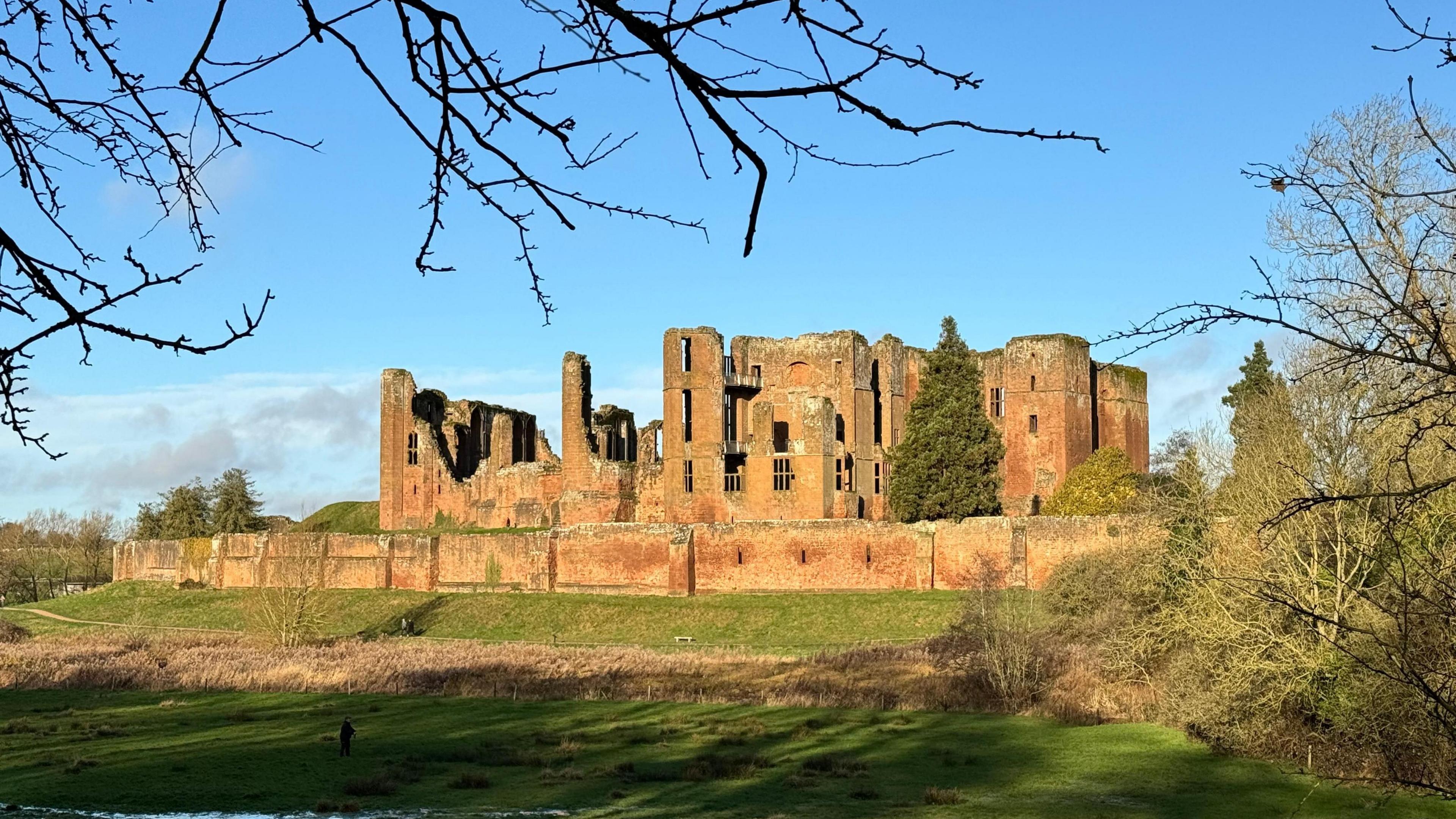 A ruined castle with several walls and turrets still standing is beneath a blue sky and behind green, grassy fields with the branches of trees just appearing at the top of the photo.