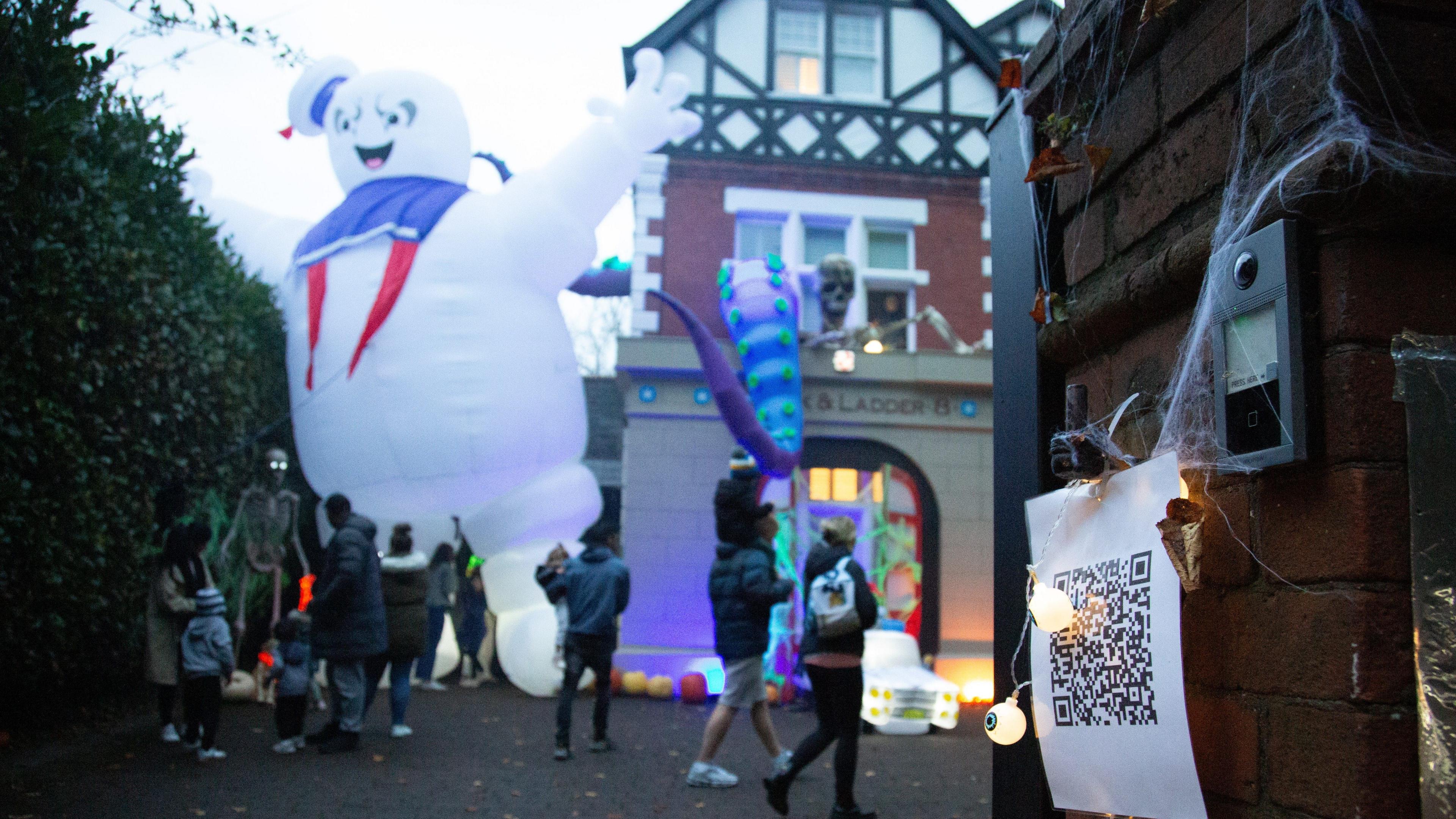 A QR code stuck to the wall outside the house with artificial cobwebs around it. The Halloween house can be seen in the background with people admiring the display.