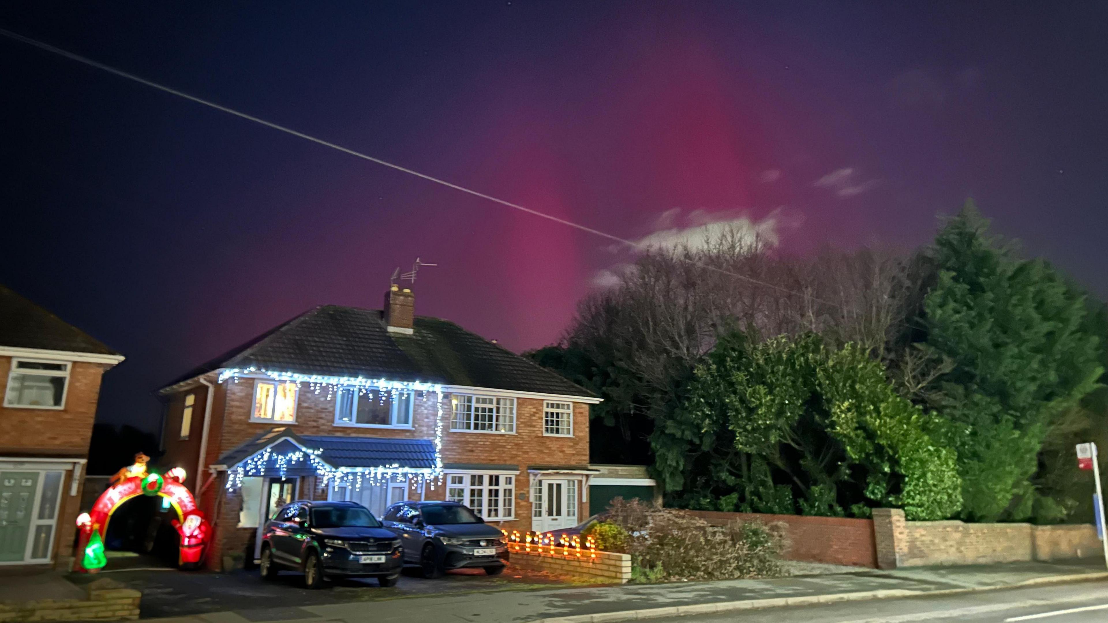 An evening sky with purple and pink hues and stars over a house decorated with Christmas lights and two black cars parked in front. Three evergreen trees are to the right of the photo 