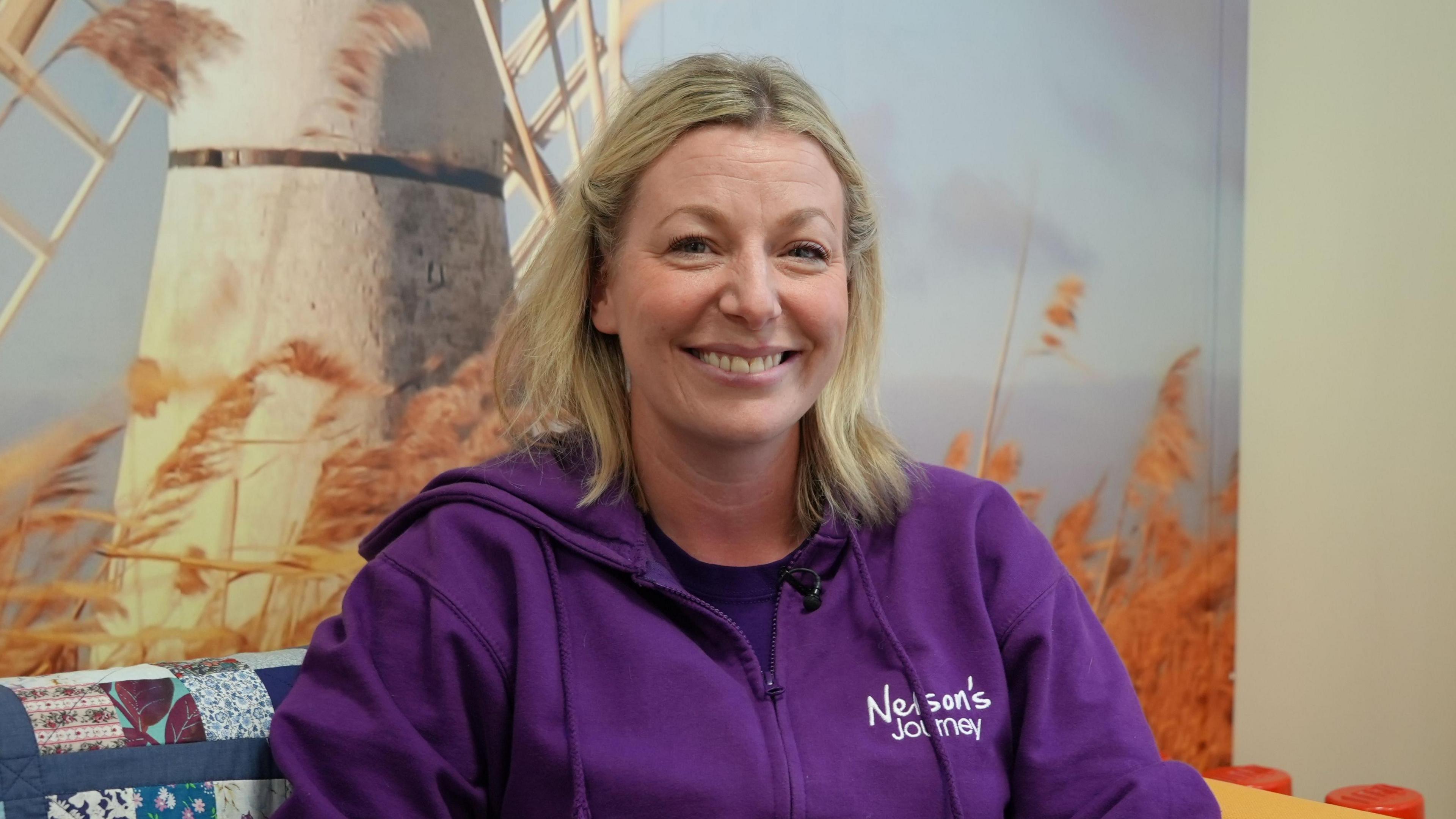 Woman sitting on a mustard sofa while smiling to camera with a picture of a windmill on the wall behind her.  She has blonde shoulder length hair and is wearing a purple zipped hoodie with the word Nelson's Journey on the breast.     