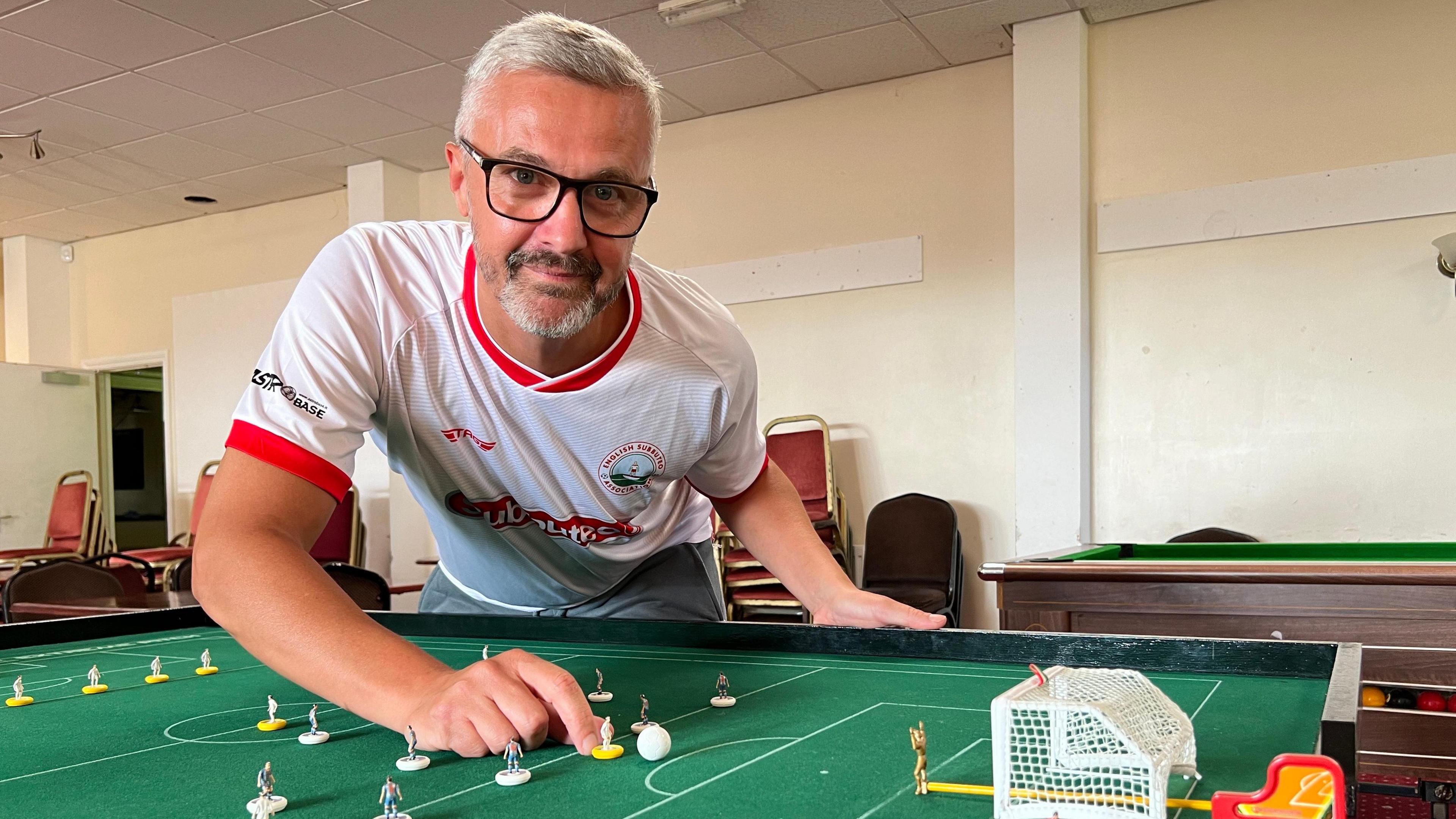 Aaron Skinner wearing white t-shirt and leaning over Subbuteo table with miniature players on.