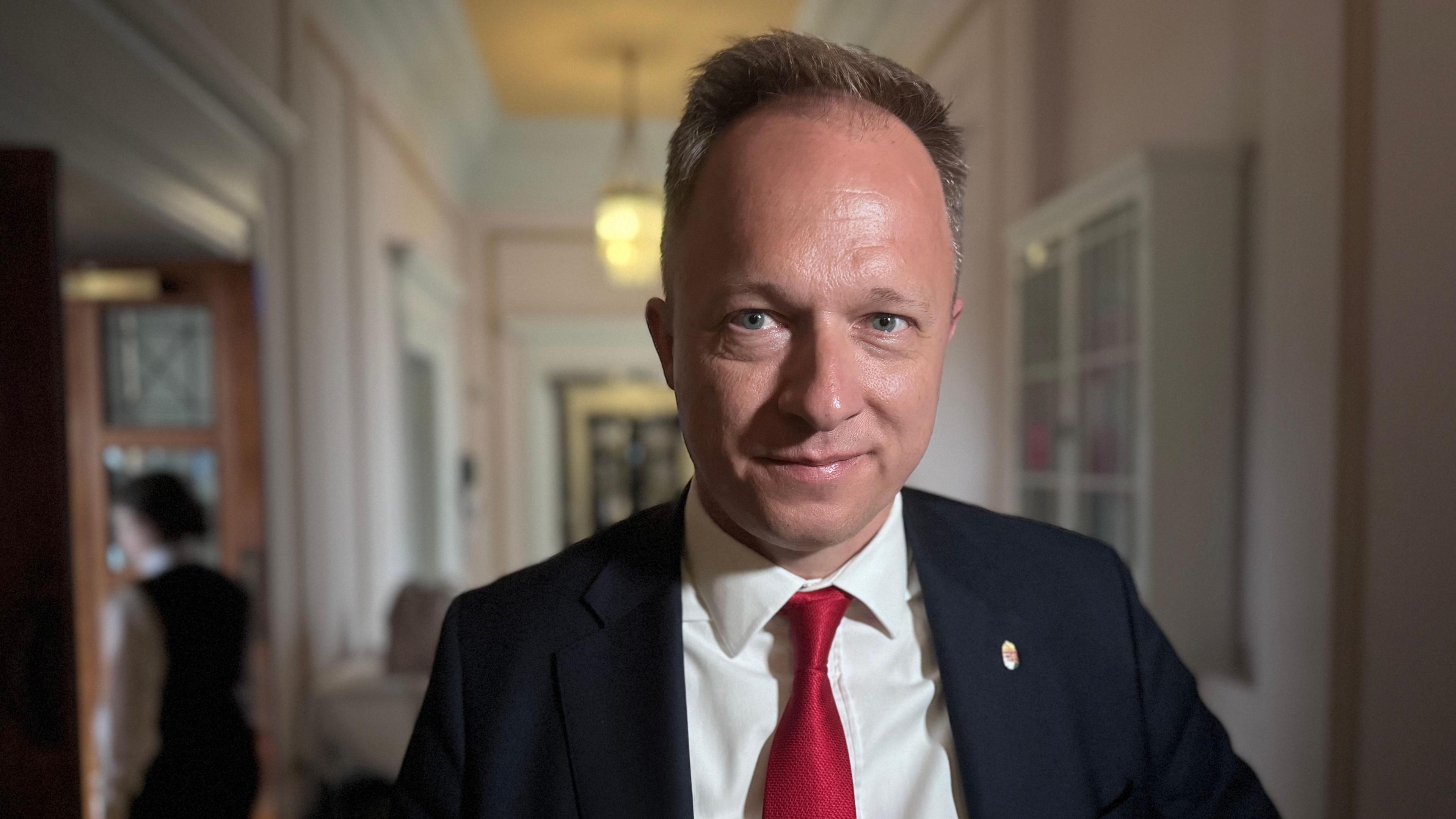 A man with brown greying hair stares into the camera. He has a black jacket, white shirt and red tie.