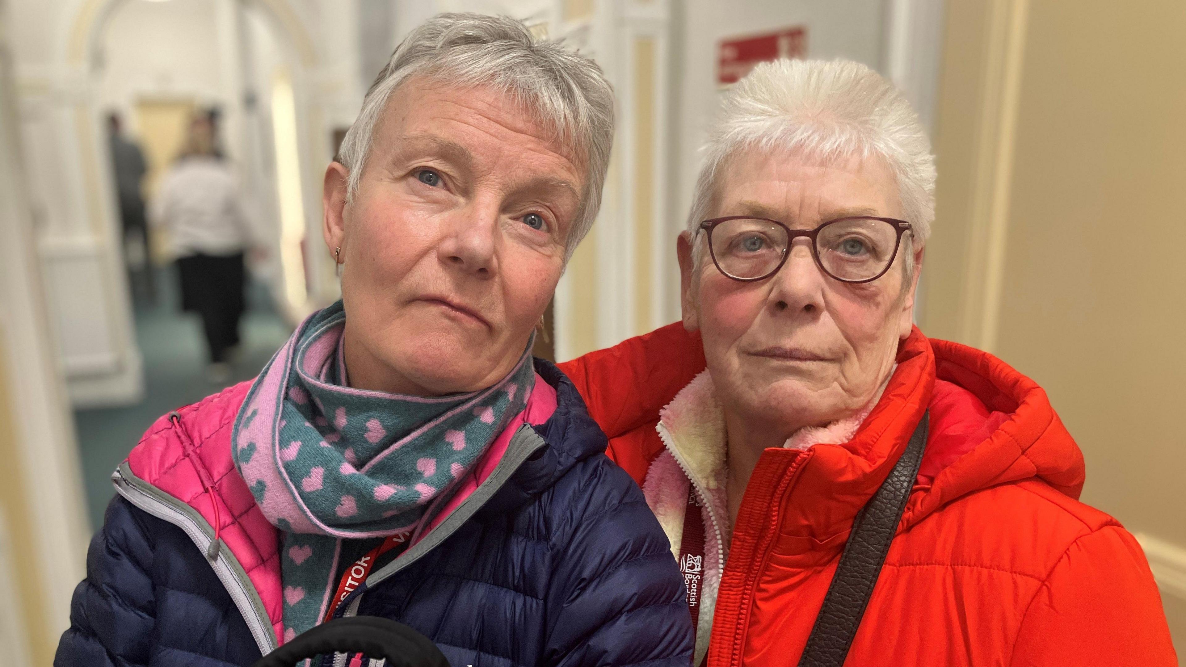 A women with grey hair in a blue jacket wearing a scarf with pink hearts stands next to another woman with grey hair and glasses in a red jacket