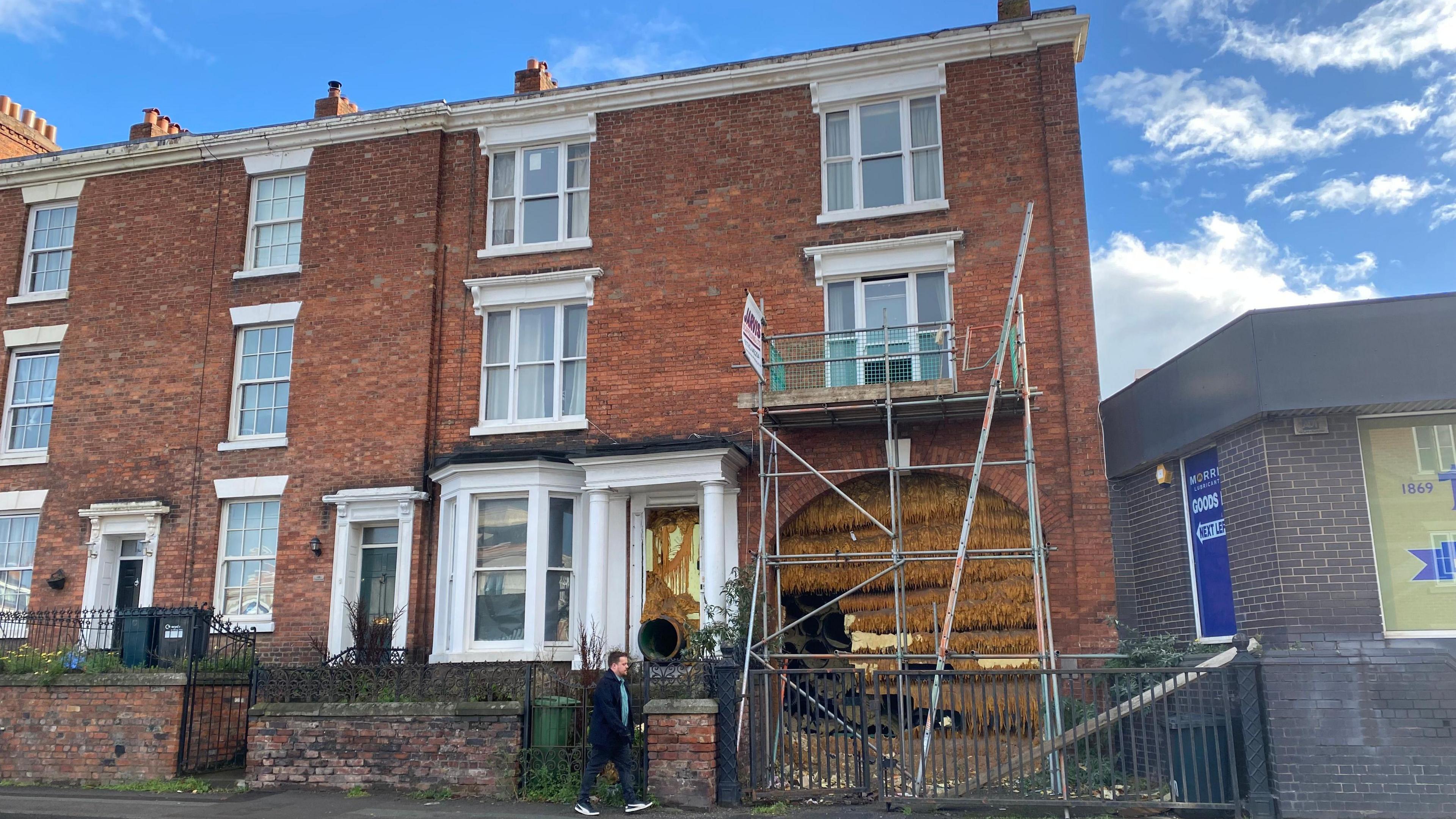 A large brown brick building that is three storeys with a large archway. The archway and doorway are filled with dark yellow solidified foam, and large tubes. There is scaffolding in front of the archway.