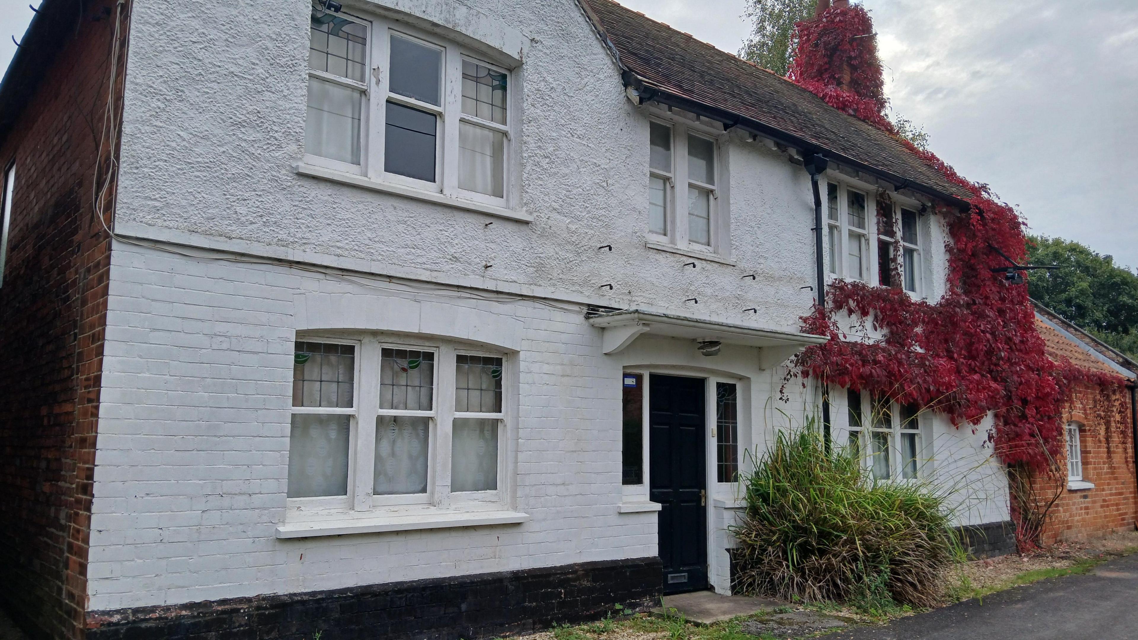 A white-painted brick-built pub, with a black door and five windows. The leaves of a red climbing plant cover one end of the building.