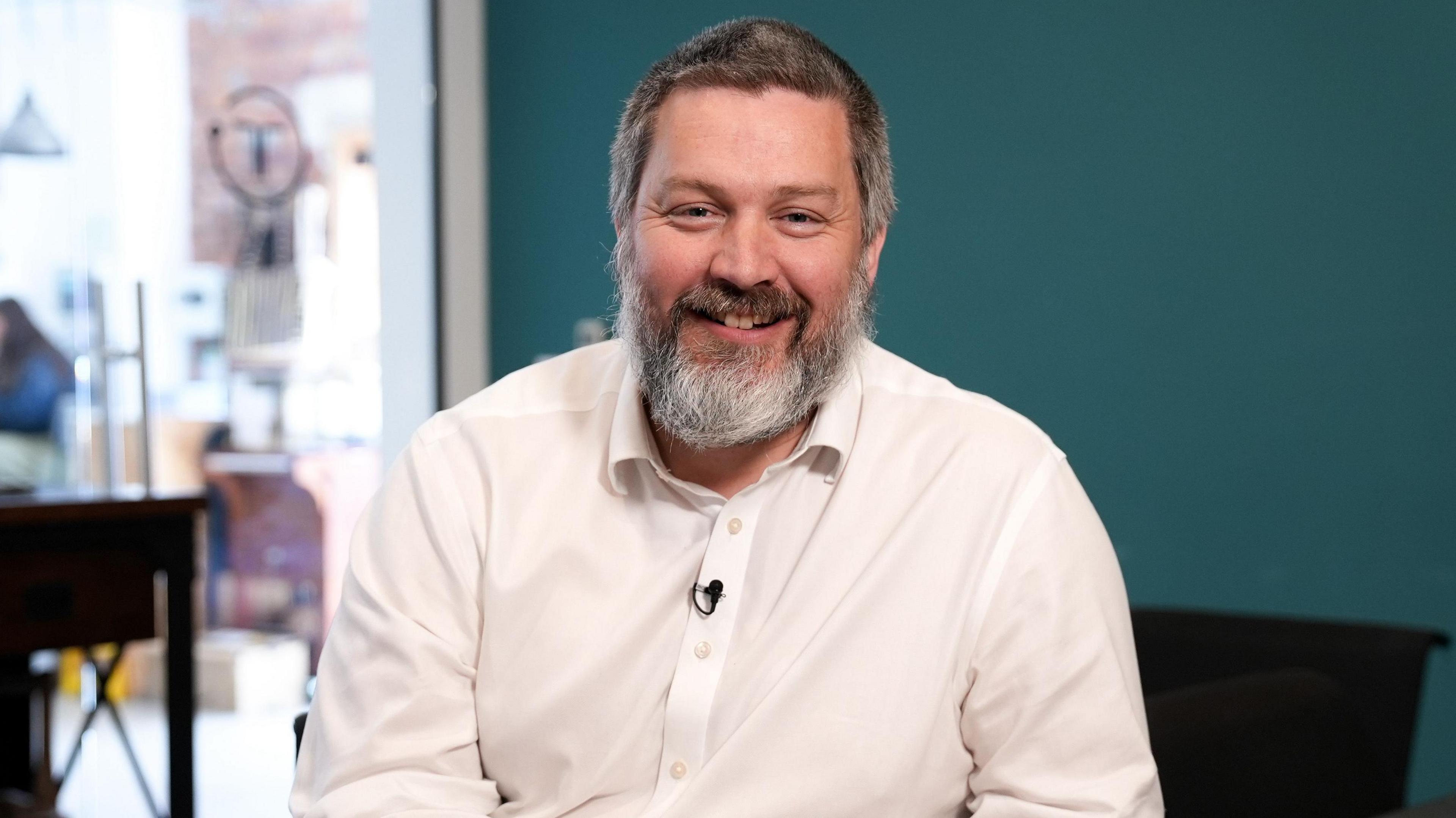 Charles Radclyffe in a medium close-up shot, sitting at a table with a teal-coloured wall across most of the background. He has a beard and his hair is dark and greying, he wears a white shirt and is smiling at the camera.