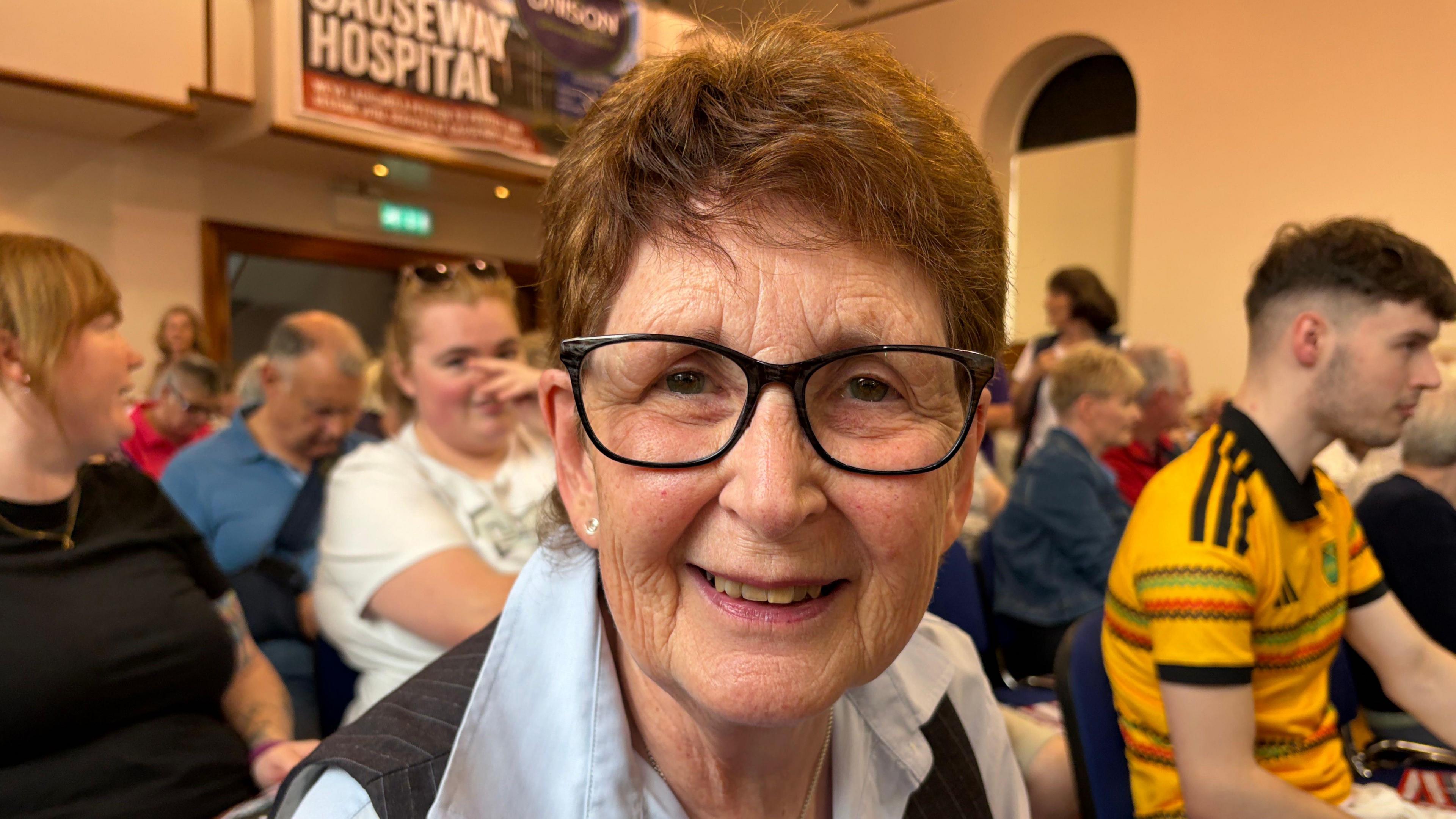 Carol Wallace has short brown hair and black glasses, she is wearing a grey pinstripe waistcoat over a light blue blouse. She is sitting in a busy meeting in Coleraine Town Hall.