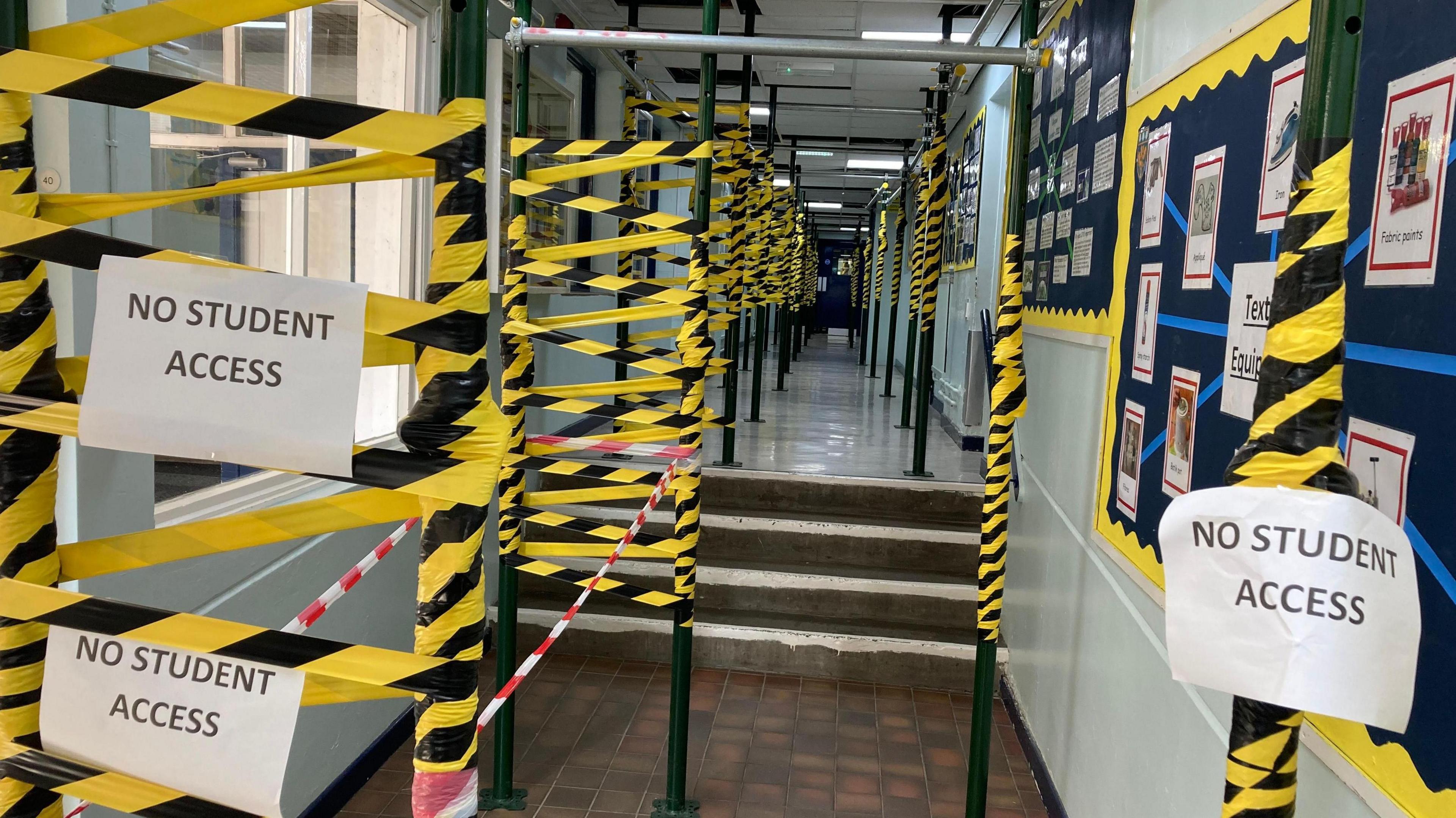 A school corridor is filled with scaffolding and vast amounts of yellow and black tape, with signs reading "no student access".