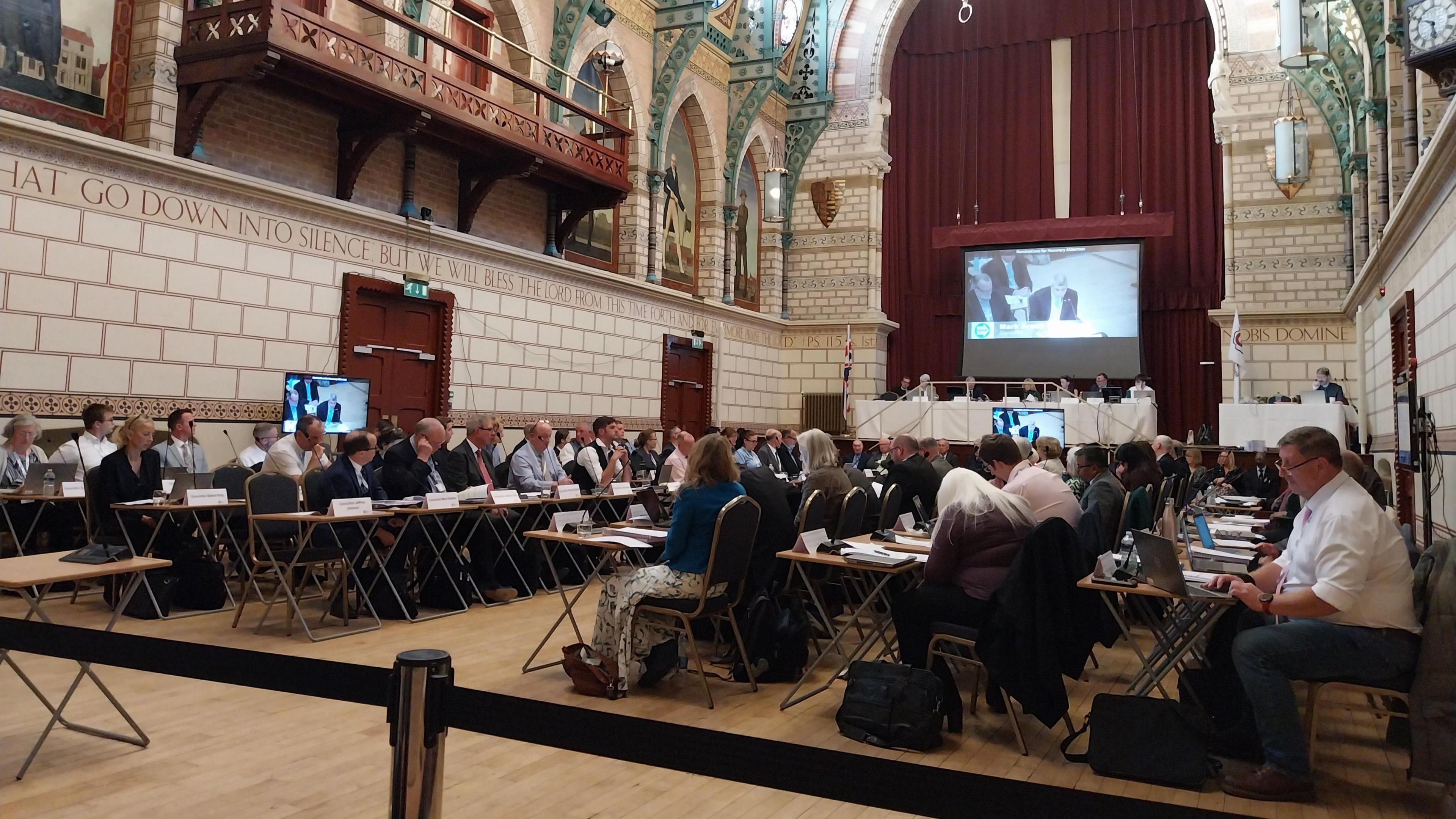 A large group of councillors in a room, holding a meeting, with three screens around them. They are sat at chairs and tables, many looking down, or at the screens. 