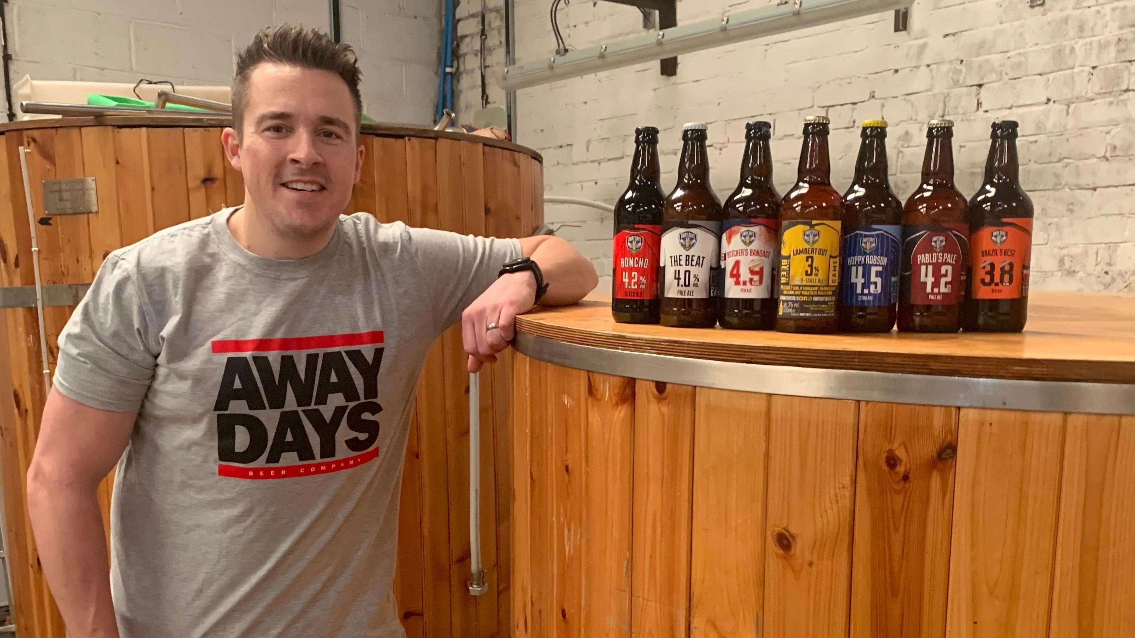 Josh Bartlett stands in a brewery with several different beer bottles placed on a brewing tank next to him. He has short dark hair and is wearing a grey t-shirt with 'Away Days' printed on it.