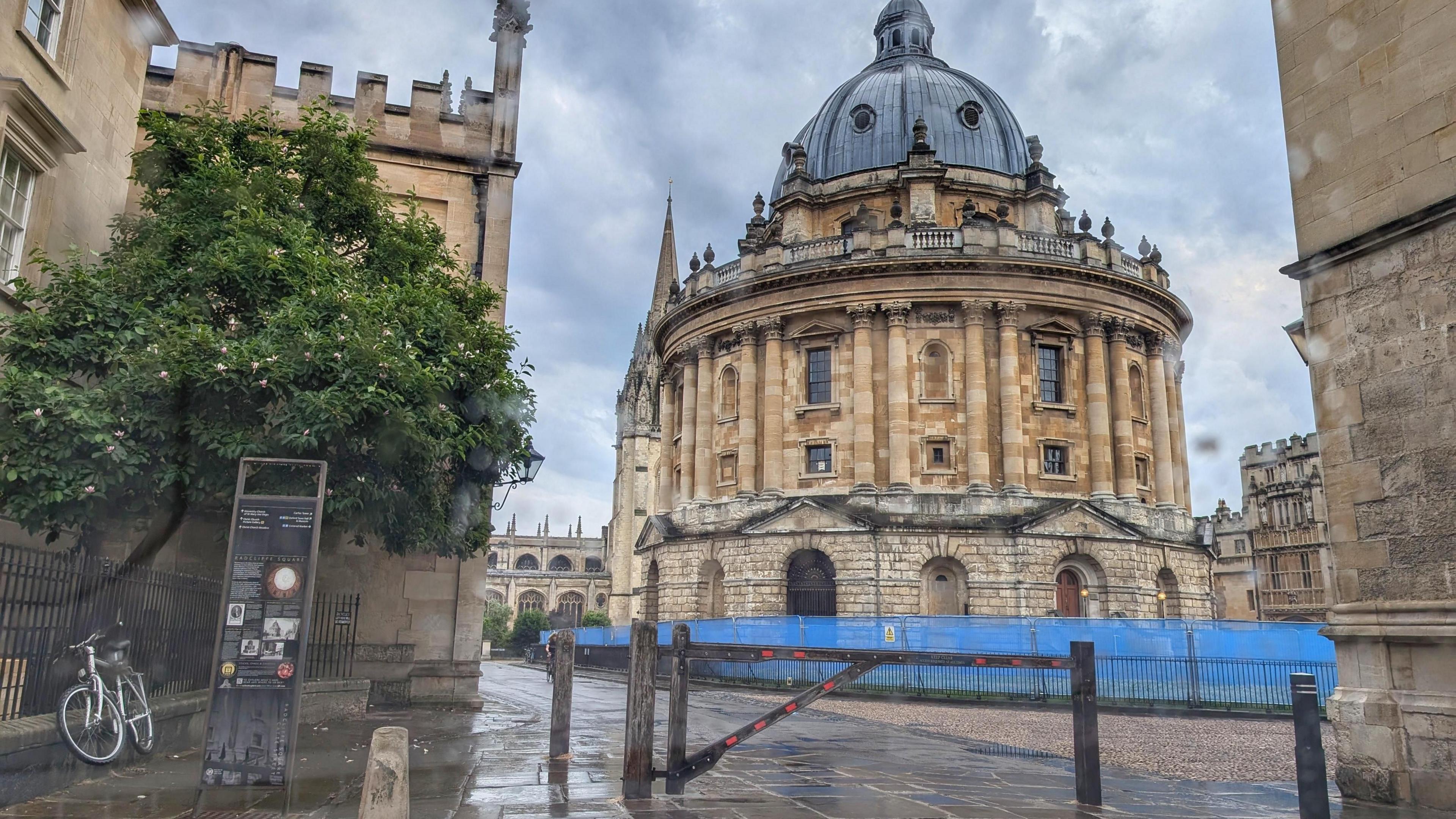 The Radcliffe Camera landmark in Oxford with rain covered pavements in front and a grey sky filled with clouds above