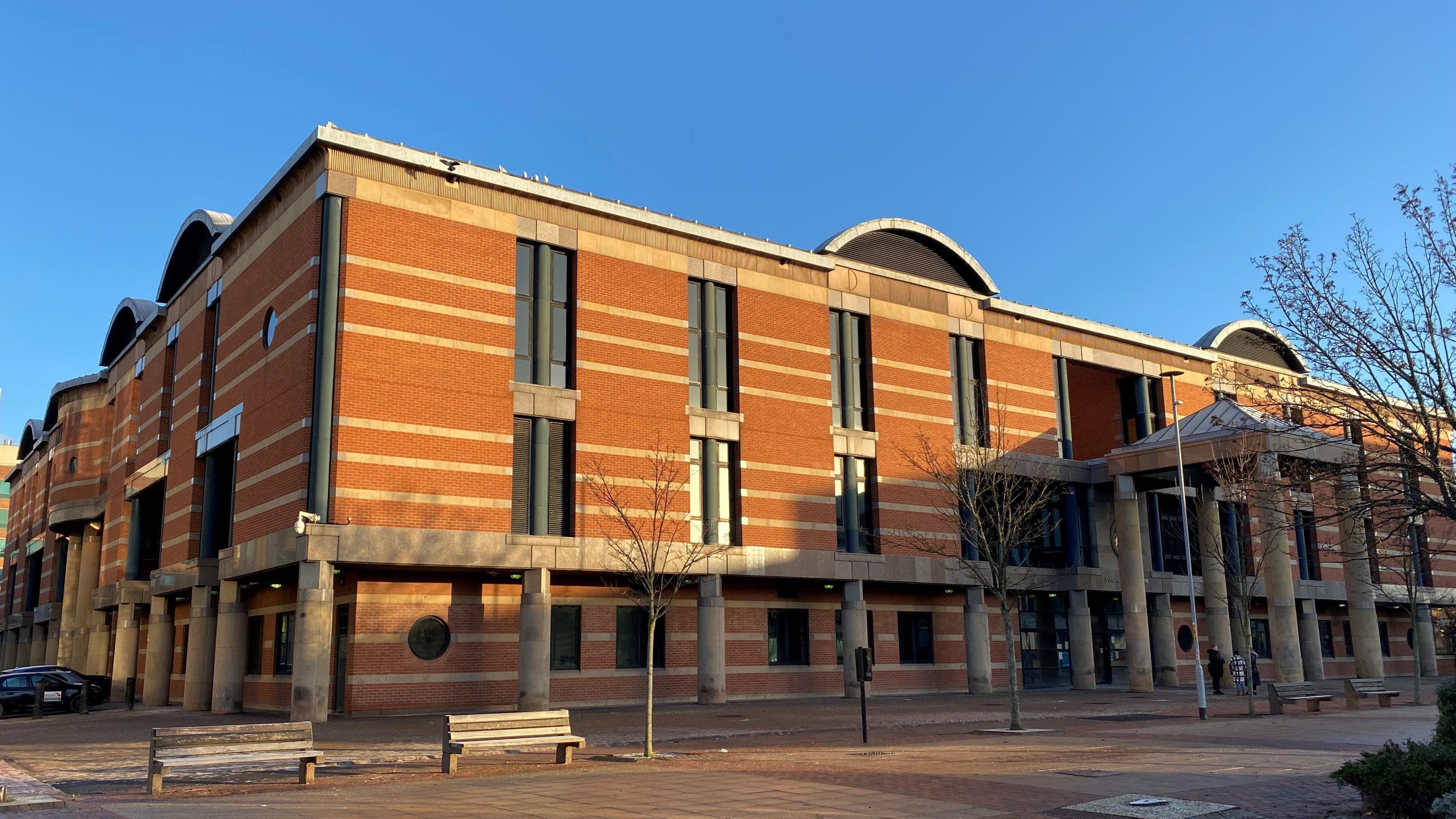 A red brick building with tall narrow windows with the sun shining on it and a blue sky beyond.